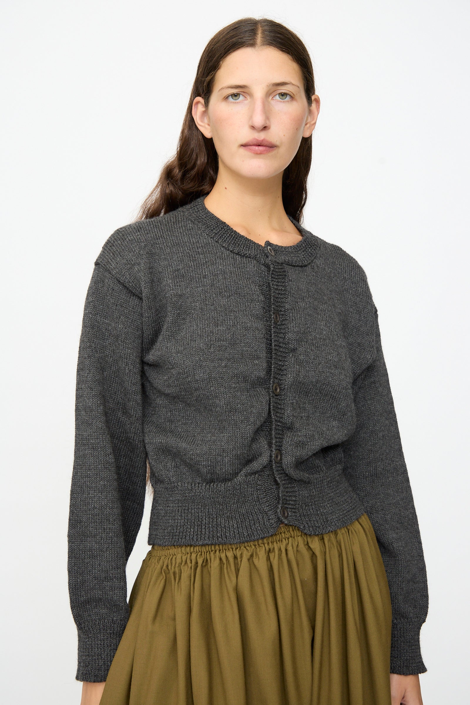 A woman stands against a plain white background wearing the SONO Caspa Cardigan in Charcoal—a dark gray button-up crewneck crafted from a luxurious wool-alpaca blend and made in the UK—paired with an olive green skirt.
