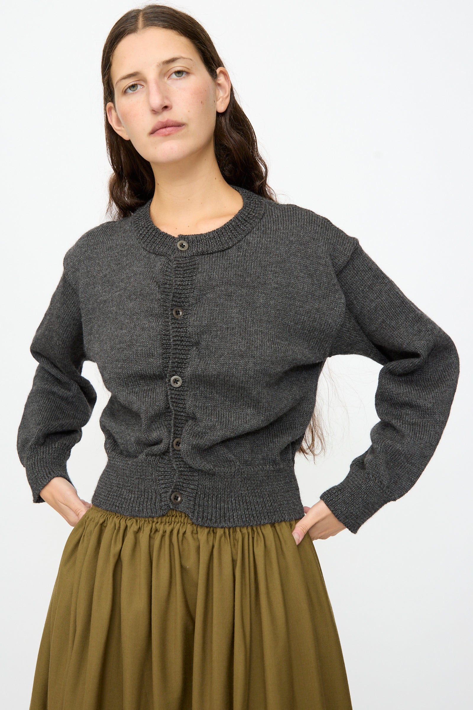 A woman stands with her hands on her hips, wearing the SONO Caspa Cardigan in Charcoal and an olive green skirt against a plain white background.