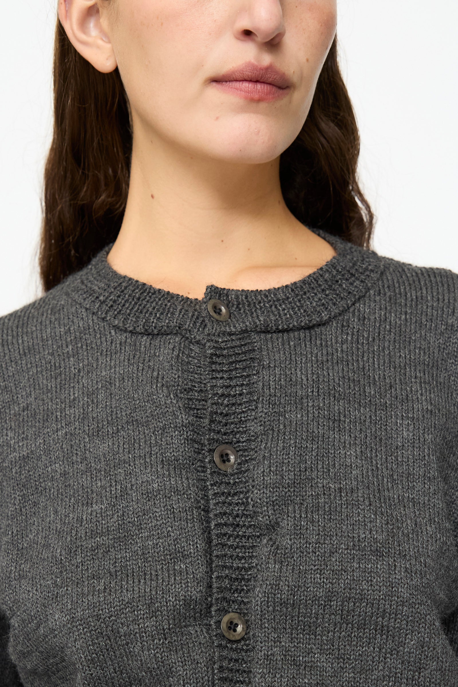A woman is shown from the upper chest to just above the lips, wearing the SONO Caspa Cardigan in Charcoal—a dark gray wool alpaca crewneck cardigan made in the UK—against a plain background.