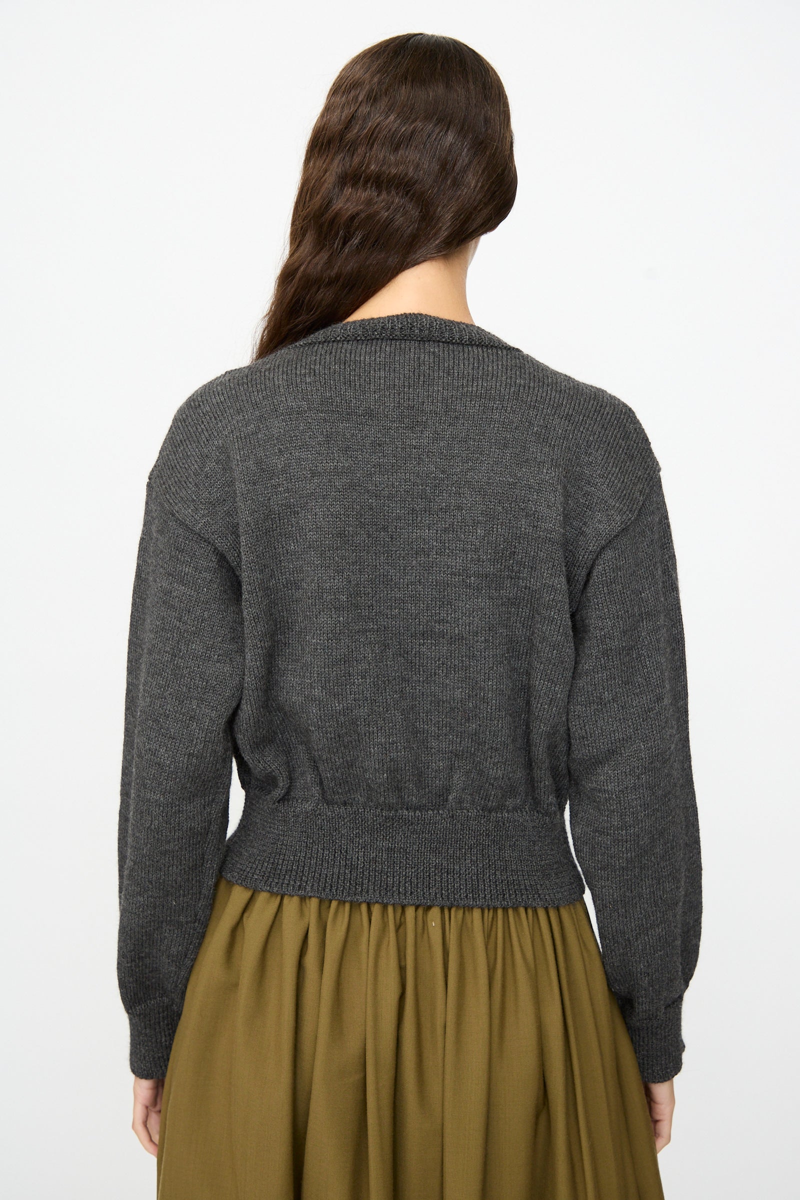 A person with long brown hair is shown from the back against a plain light background, wearing the SONO Caspa Cardigan in Charcoal and an olive green skirt.