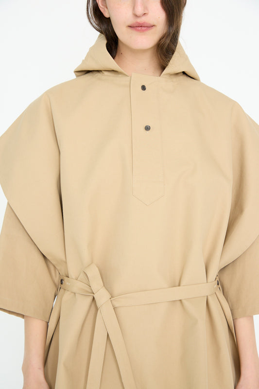 A person wears the SONO Cotton Cole Cape in Jute, featuring a beige hooded design made from organic cotton with button closure and waist tie, against a plain background.