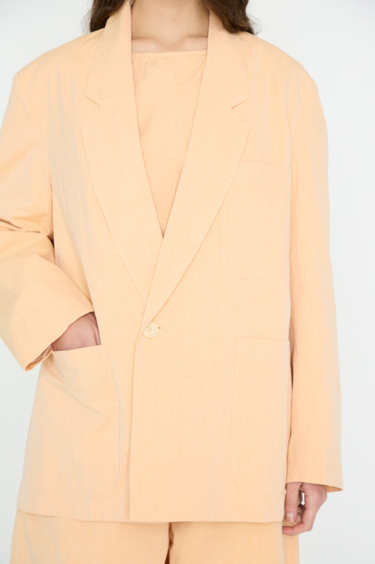 Against a plain background, the person wears SONO's Cotton Juri Jacket in Dusty Peach, made from organic cotton. Their right hand is tucked into the pocket of the oversized blazer.