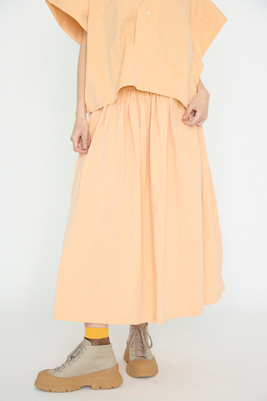 A person stands against a white background, wearing a wide silhouette with a loose top and SONO's Cotton Skye Skirt in Dusty Peach, paired with beige chunky boots and yellow socks. 