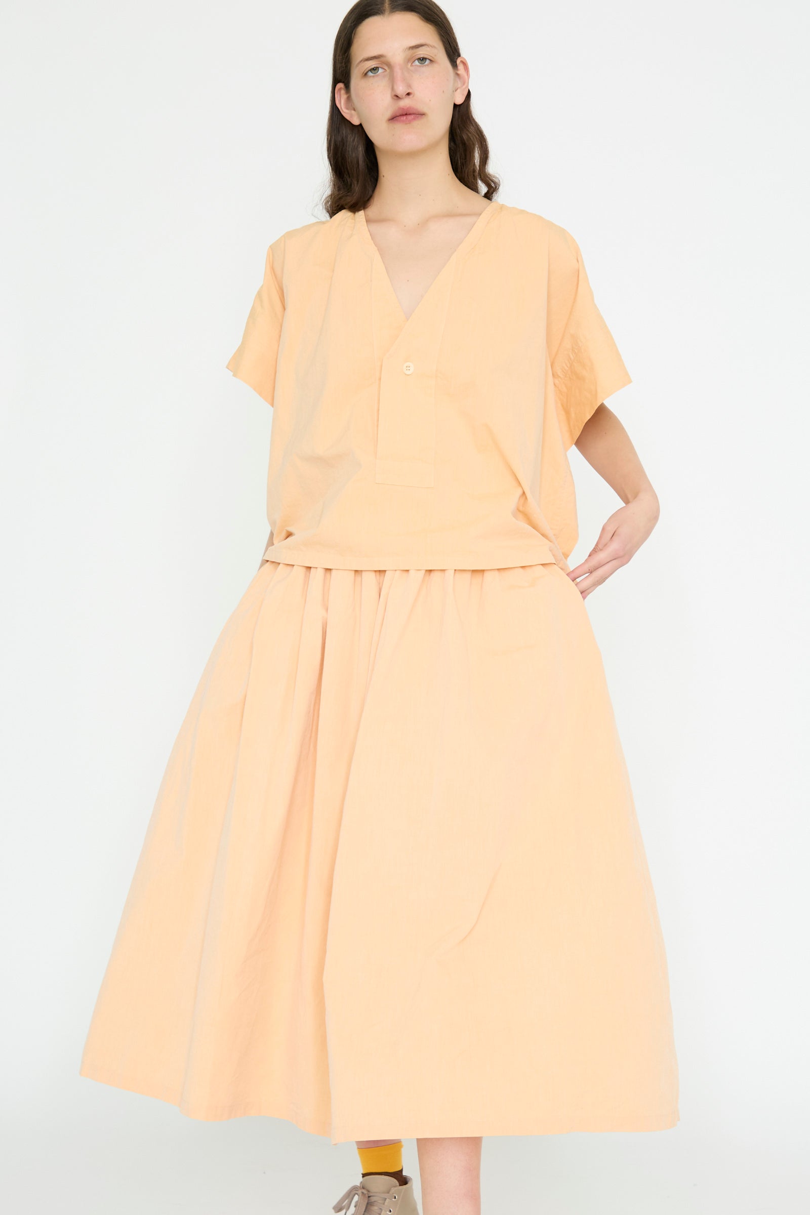 Against a white backdrop, a person is wearing the Cotton Skye Skirt in Dusty Peach by SONO and a pale orange blouse. 
