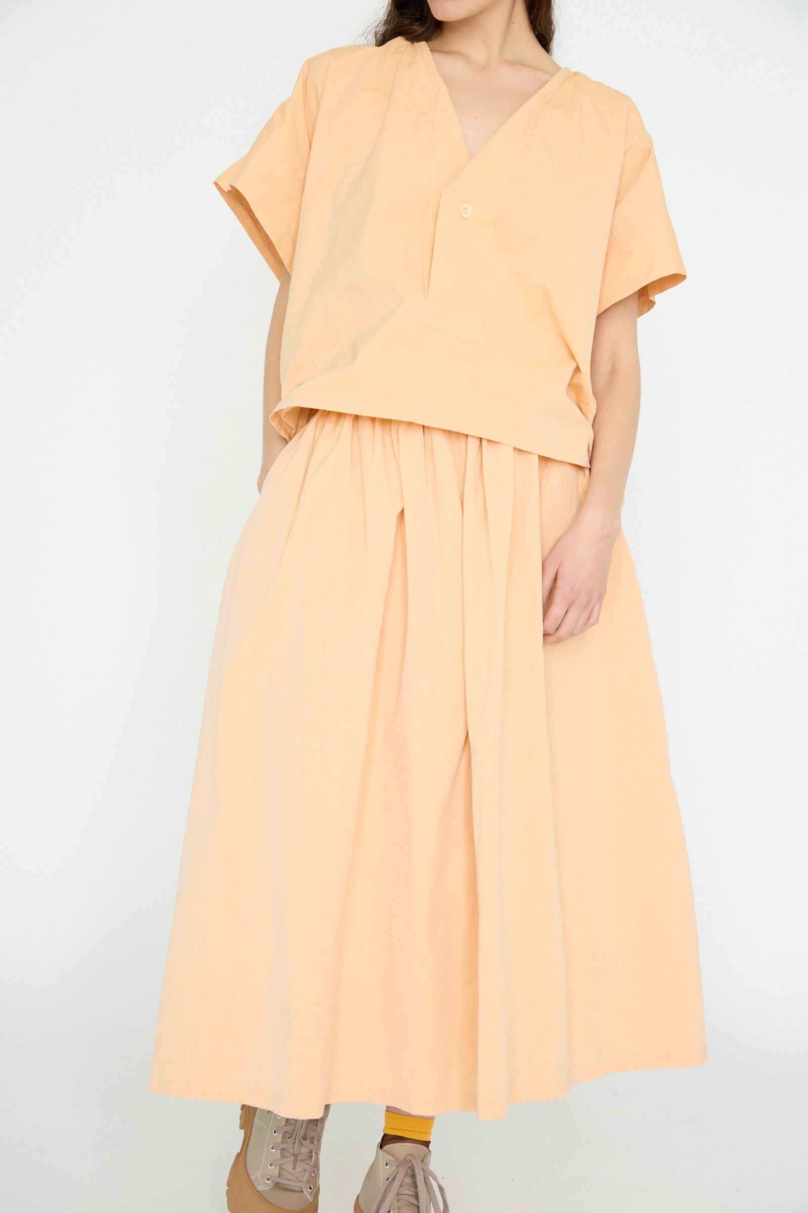 A person wears a loose-fitting top and the SONO Cotton Skye Skirt in Dusty Peach, paired with tan sneakers and yellow socks, set against a plain white backdrop. Made in London.