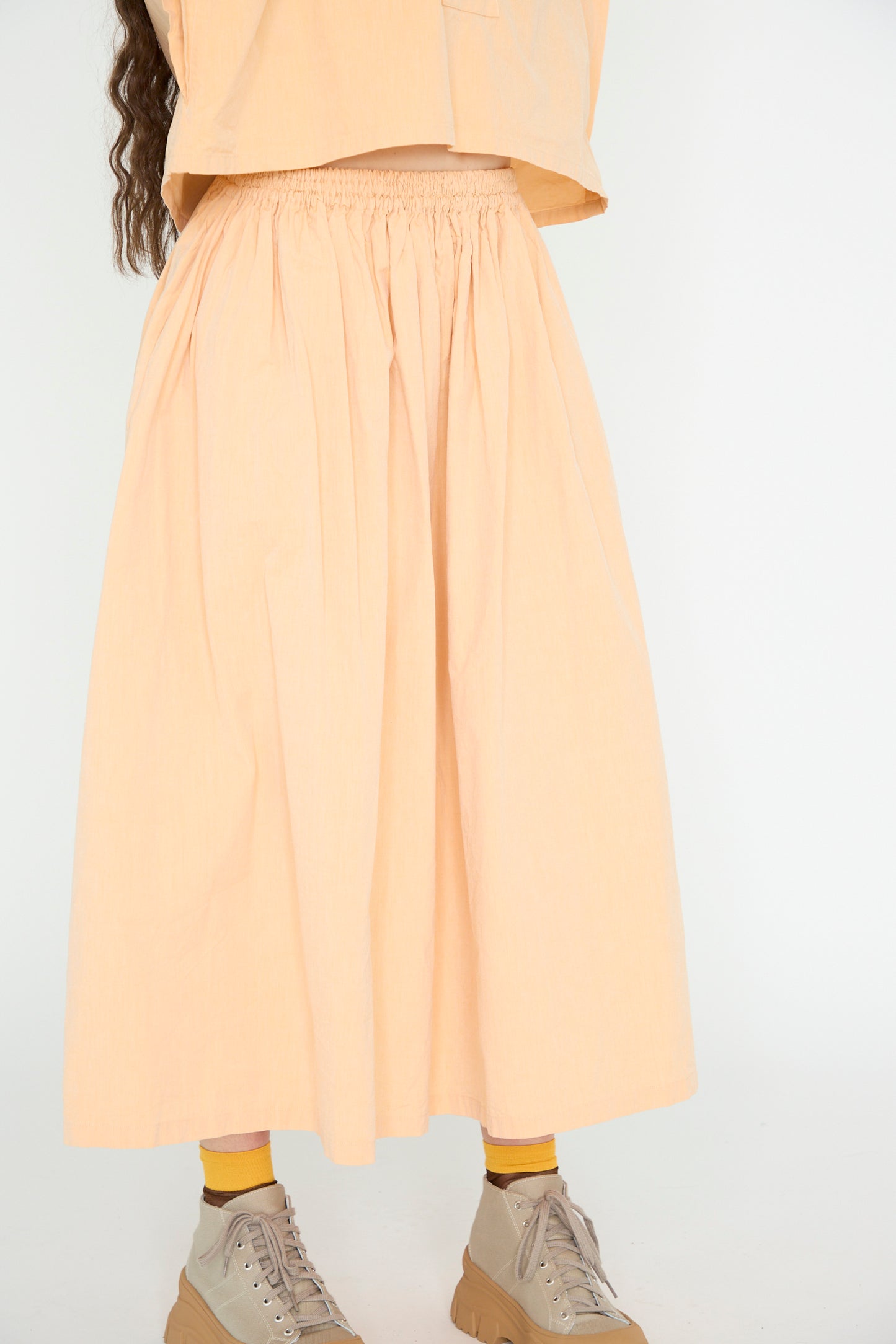 The image shows SONO's Cotton Skye Skirt in Dusty Peach, featuring a wide silhouette and an elastic waistband, worn with yellow socks and tan lace-up shoes. 