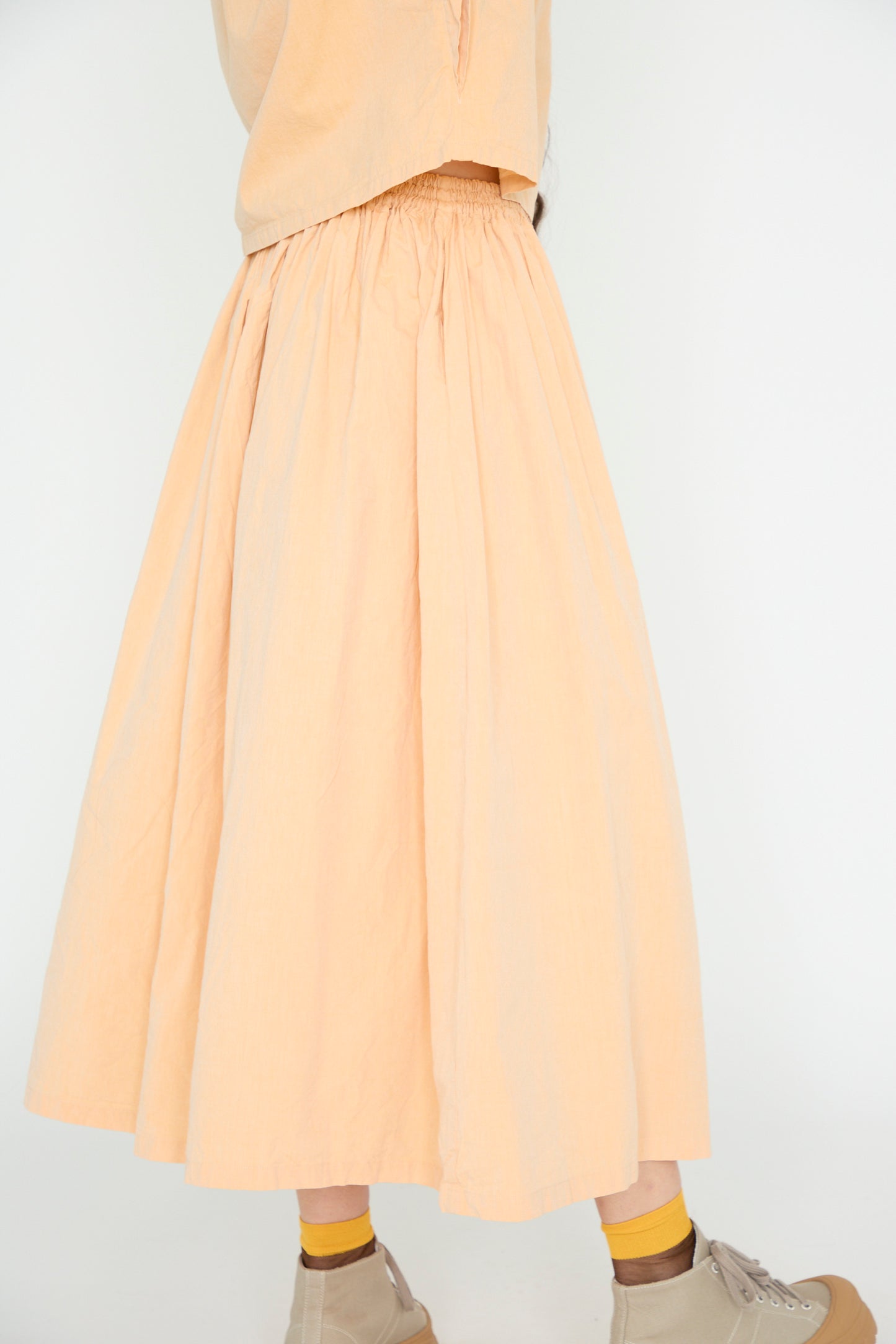 A person wearing SONO's Cotton Skye Skirt in Dusty Peach paired with tan shoes and yellow socks strikes a wide silhouette against a plain white background.