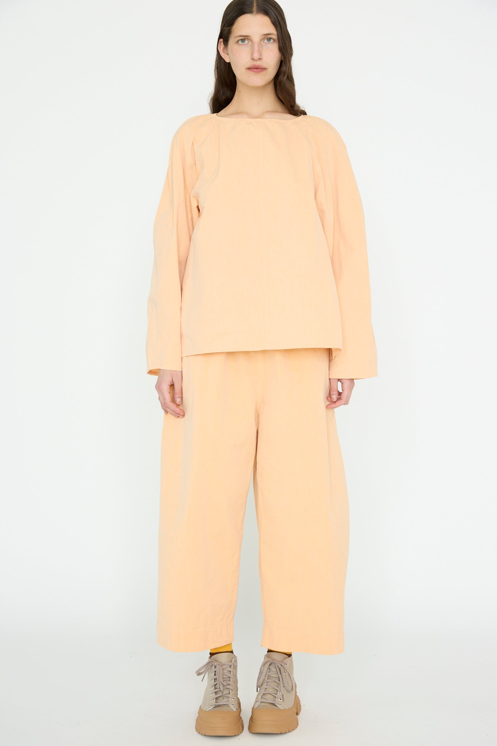 A person stands against a white background wearing the SONO Cotton Tucker Trouser in Dusty Peach, a loose-fit with wide cropped legs, complemented by beige shoes.