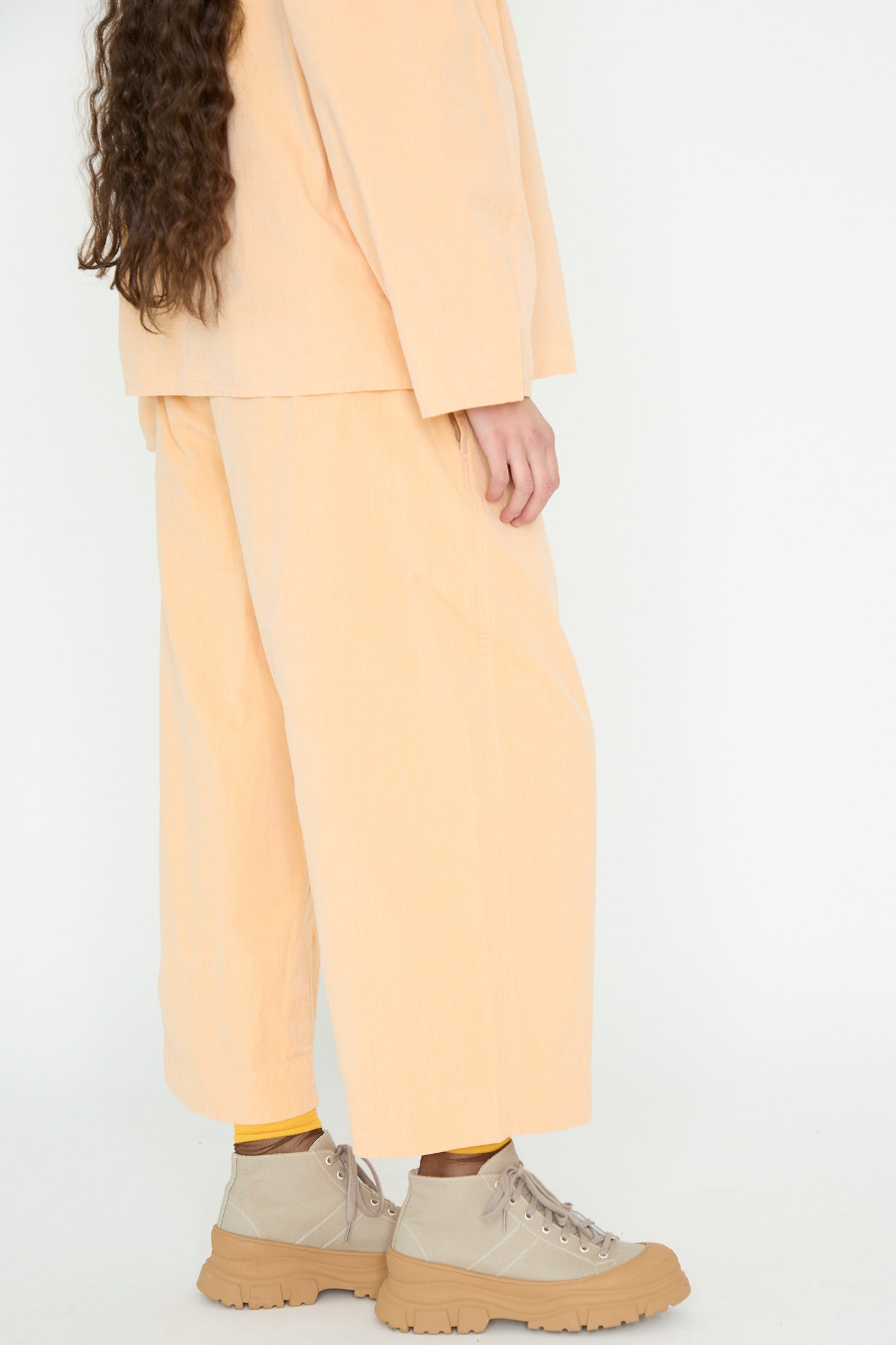 Dressed in SONO's Cotton Tucker Trouser in Dusty Peach with wide, cropped legs and long sleeves, the ensemble is completed with beige high-top sneakers.