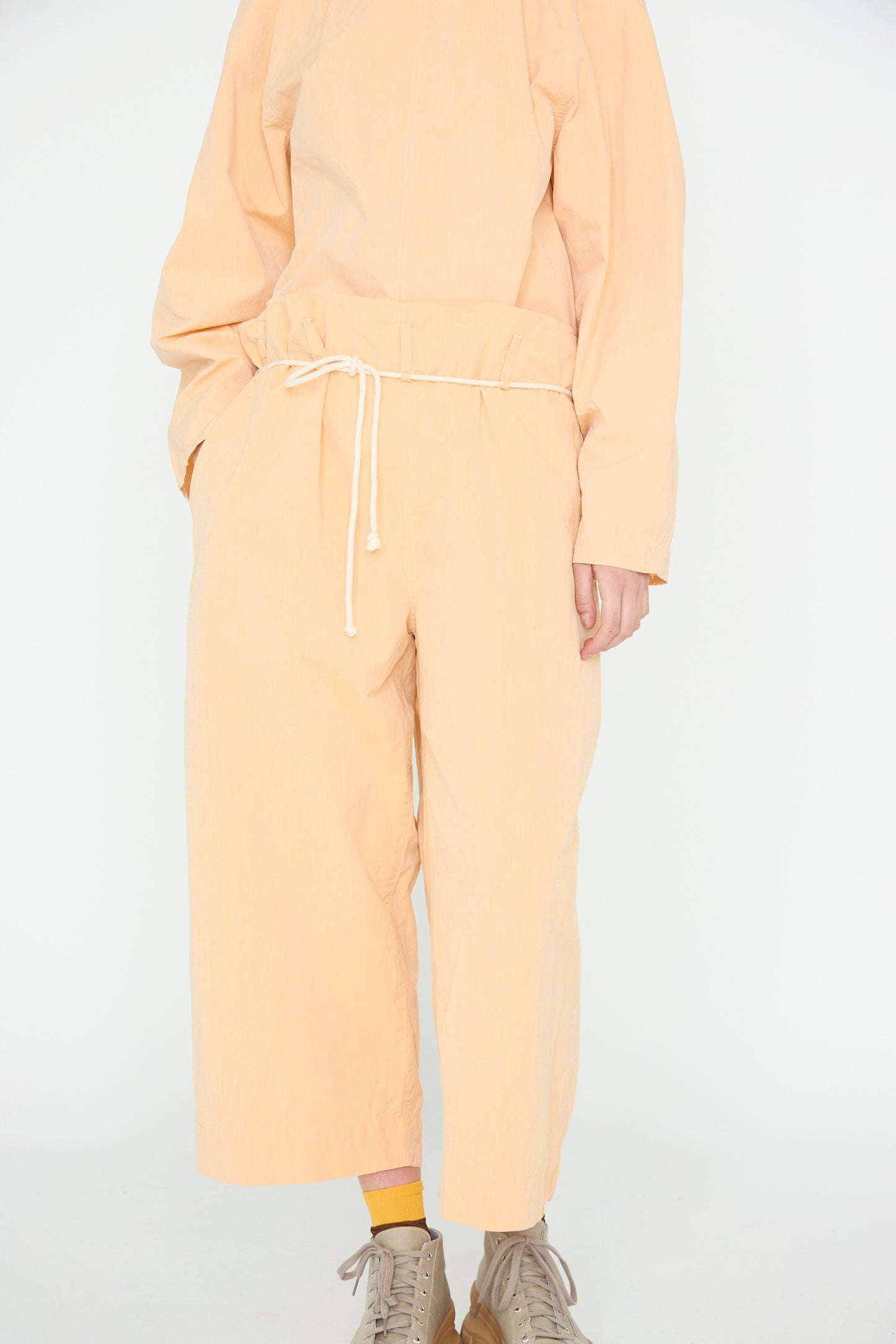 Someone wears SONO's Cotton Tucker Trouser in Dusty Peach, featuring a drawstring waist and wide cropped legs. The outfit is paired with beige shoes and vibrant yellow socks, against a plain white backdrop.
