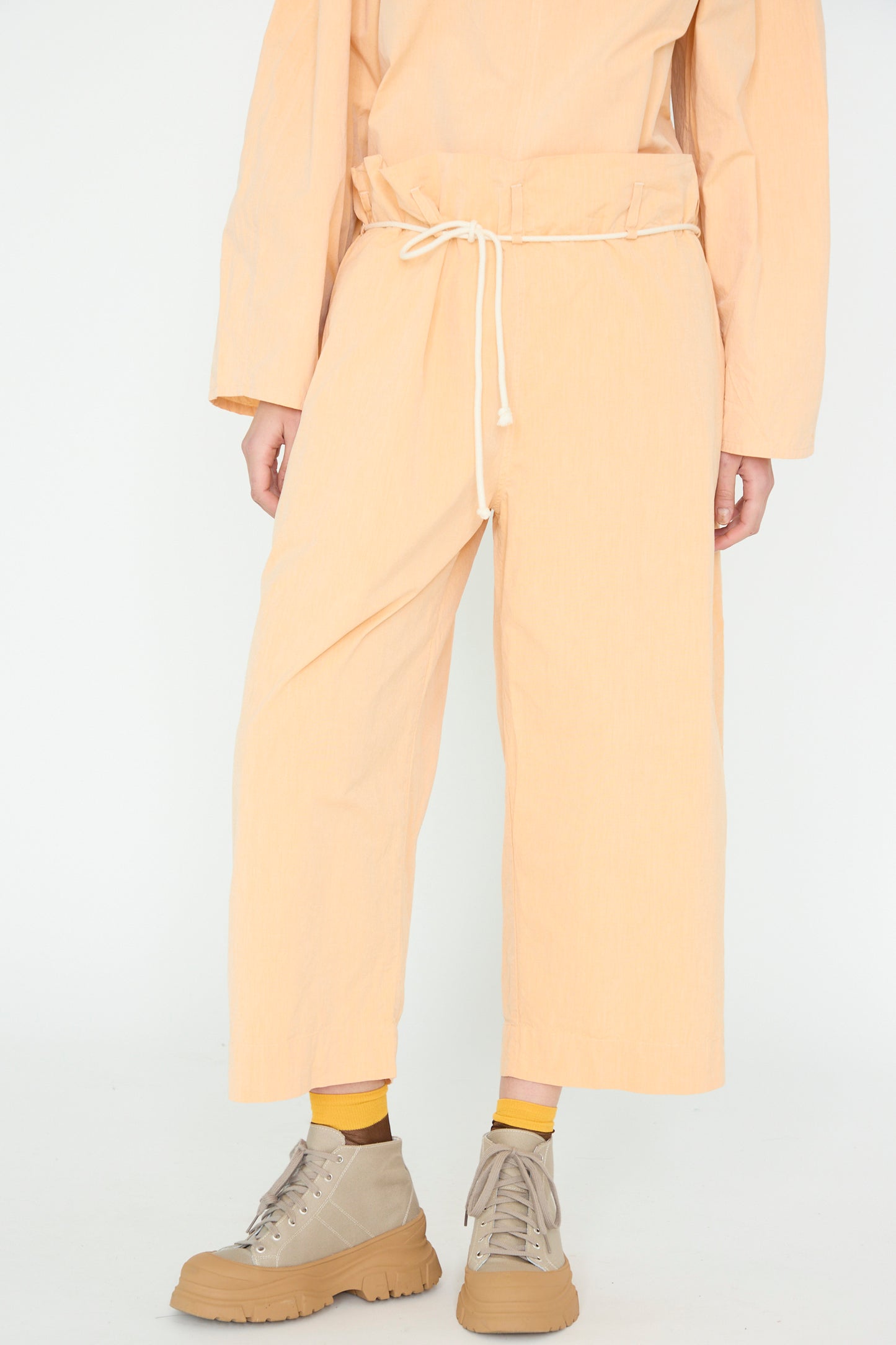 Wearing SONO's Cotton Tucker Trouser in Dusty Peach, the person blends organic cotton wide cropped trousers and a matching top with beige high-top sneakers and vibrant yellow socks, standing against a plain backdrop.