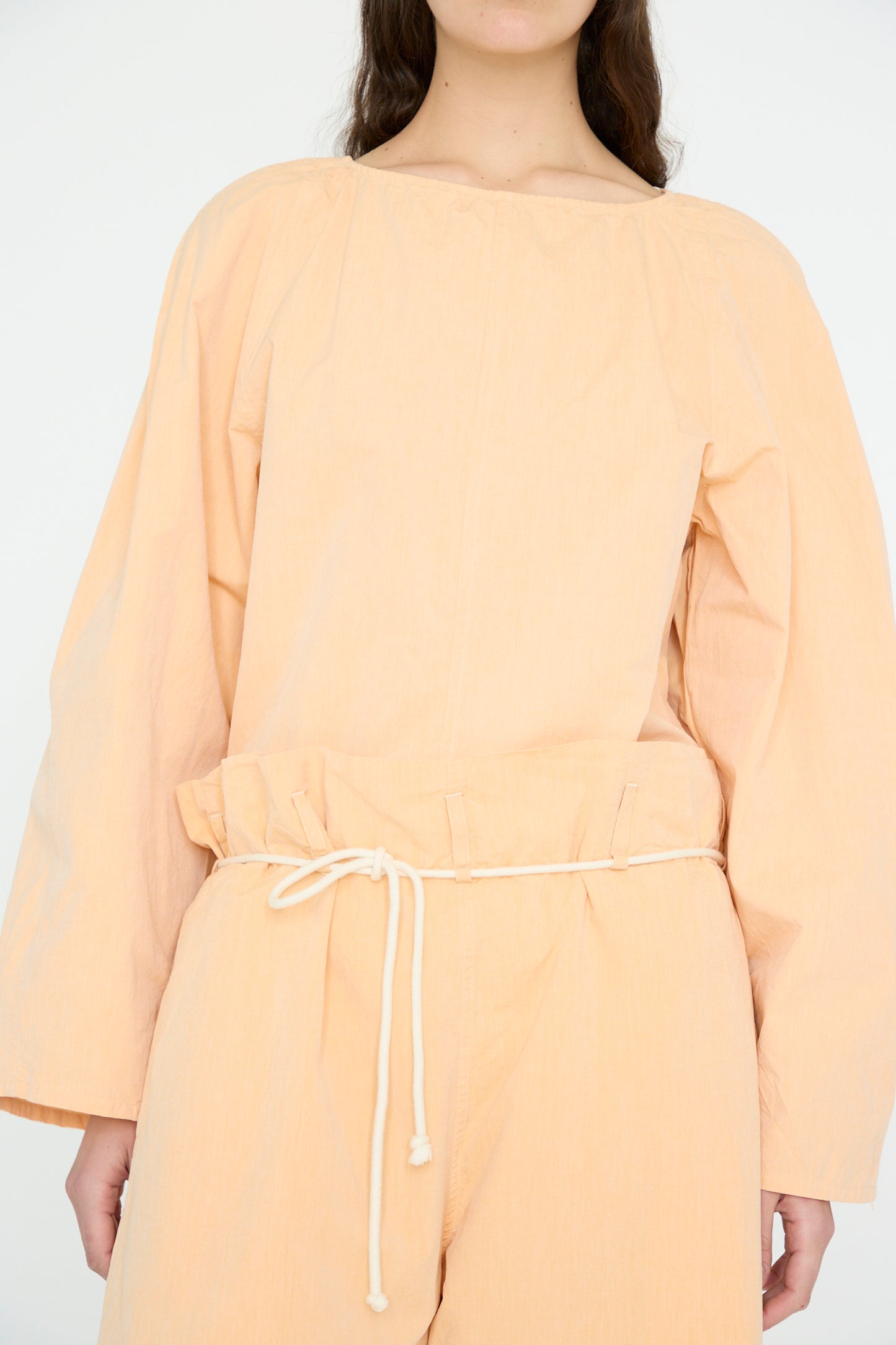 Against a plain background, a person wears SONO's Cotton Tucker Trouser in Dusty Peach with long sleeves and a drawstring belt, complemented by organic cotton trousers with wide cropped legs.