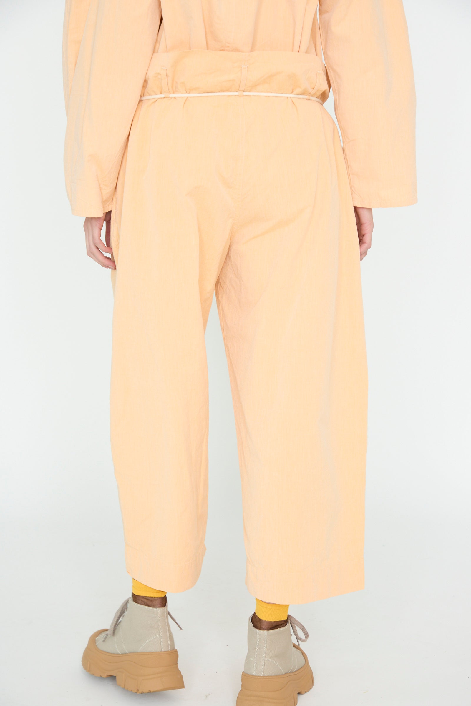 A person is photographed from behind in SONO's Cotton Tucker Trouser in Dusty Peach. They wear wide cropped leg pants made of organic cotton, paired with yellow socks and beige shoes, against a plain background.