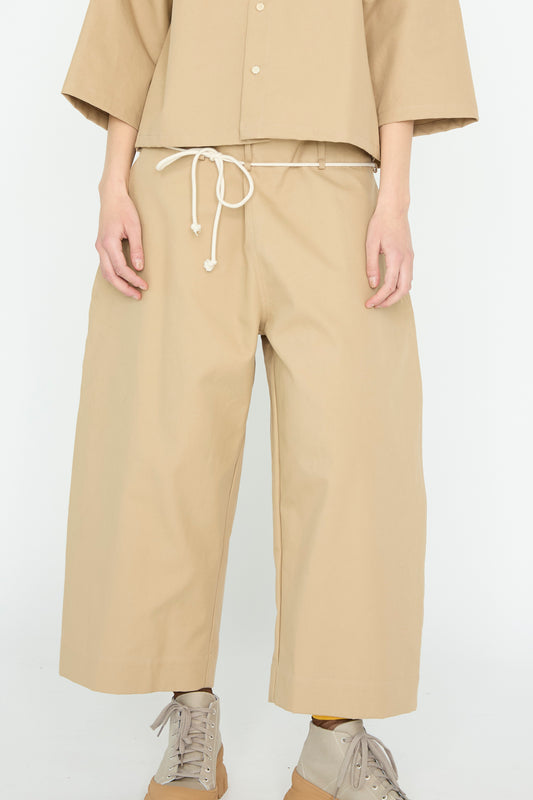 A person wears the SONO Cotton Tucker Trouser in Jute, featuring a wide cropped leg with a white drawstring, made of organic cotton. They pair it with a matching top and beige sneakers against a plain background. Stylishly crafted in London.