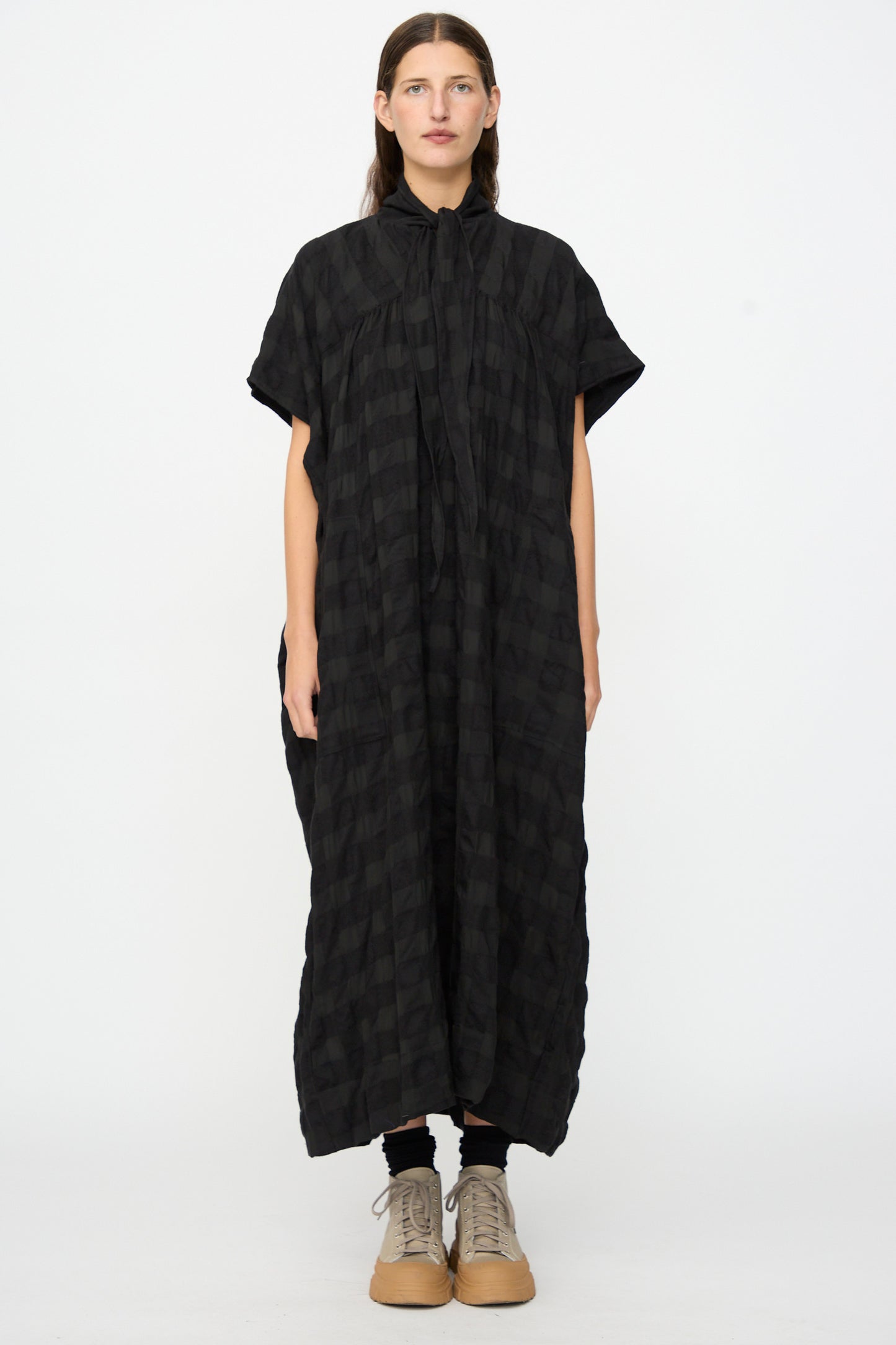 A person stands against a white background wearing the SONO Dawn Dress in Black on Black—a loose, ankle-length maxi dress with short sleeves, featuring a subtle checkered pattern and crafted from a soft wool-viscose blend—paired with tan lace-up shoes.