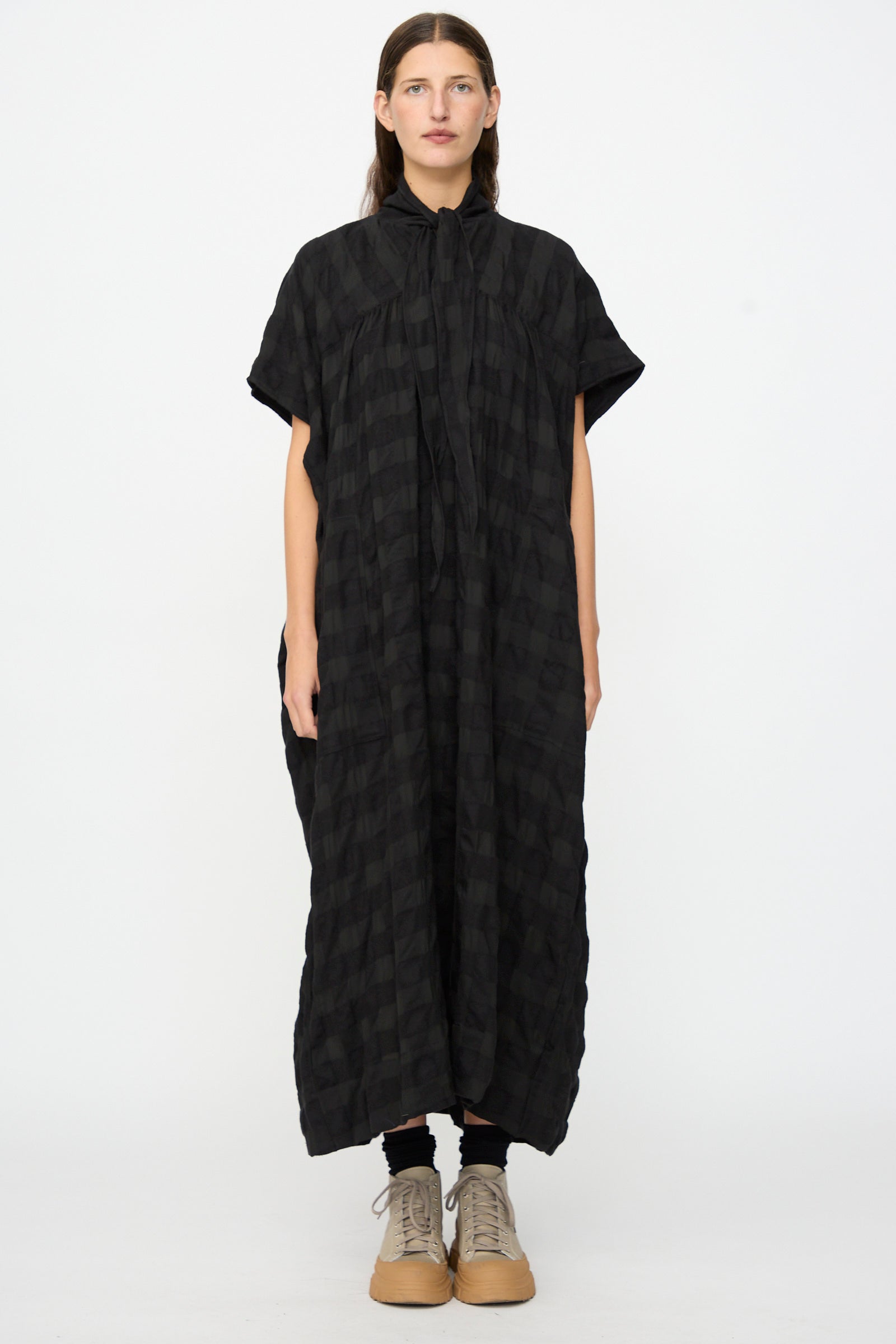 A person stands against a white background wearing the SONO Dawn Dress in Black on Black—a loose, ankle-length maxi dress with short sleeves, featuring a subtle checkered pattern and crafted from a soft wool-viscose blend—paired with tan lace-up shoes.