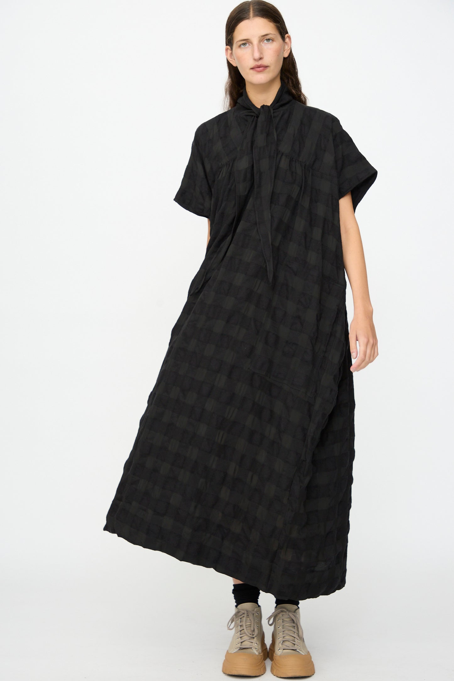 A person wearing the SONO Dawn Dress in Black on Black—a loose-fitting, short-sleeved maxi dress with a black checkered wool-viscose blend and a tie-neck detail—paired with black leggings and beige lace-up shoes, standing against a plain white background.