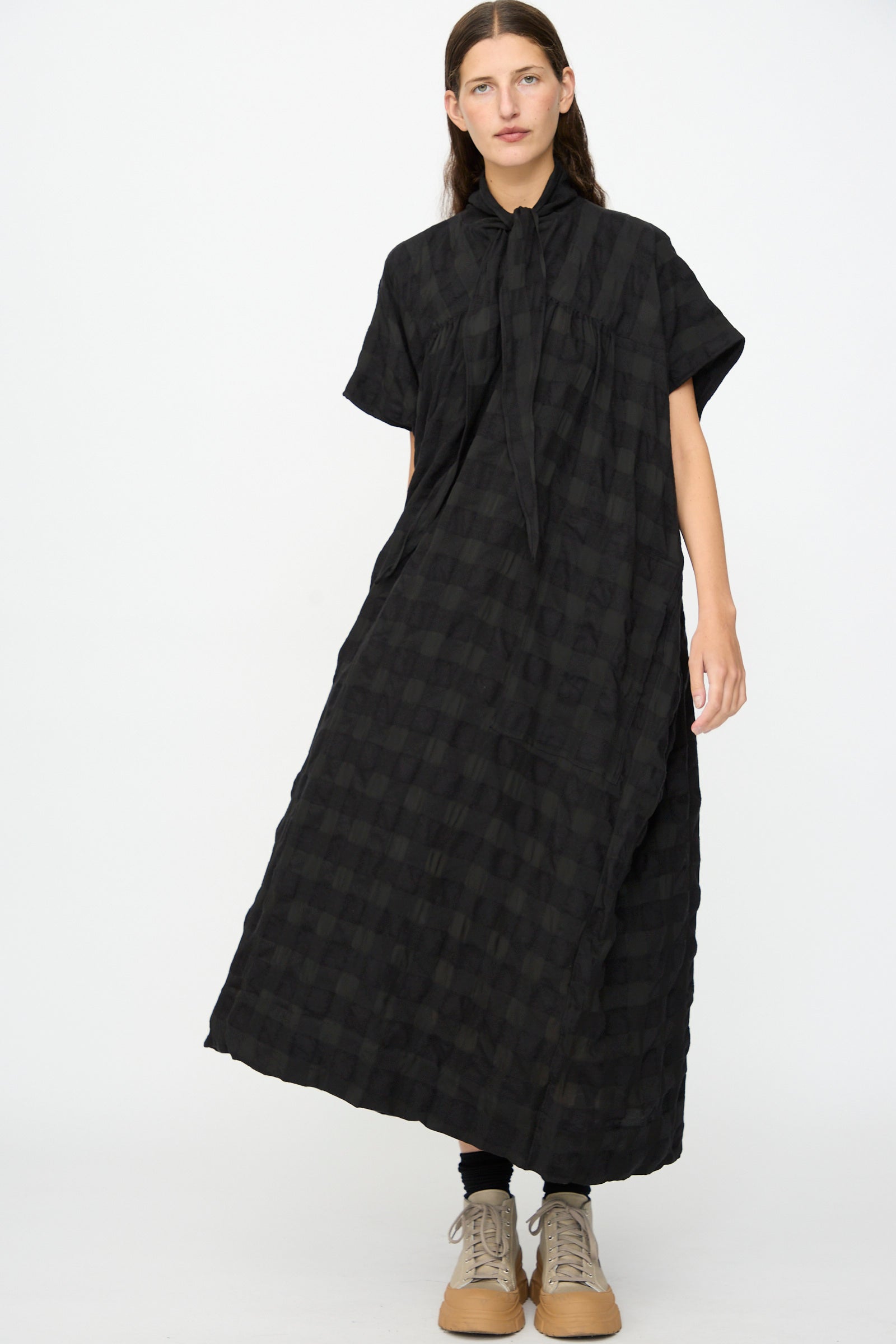 A person wearing the SONO Dawn Dress in Black on Black—a loose-fitting, short-sleeved maxi dress with a black checkered wool-viscose blend and a tie-neck detail—paired with black leggings and beige lace-up shoes, standing against a plain white background.