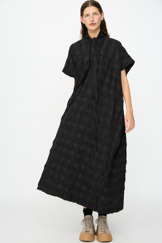 A person wearing the SONO Dawn Dress in Black on Black—a loose-fitting, short-sleeved maxi dress with a black checkered wool-viscose blend and a tie-neck detail—paired with black leggings and beige lace-up shoes, standing against a plain white background.