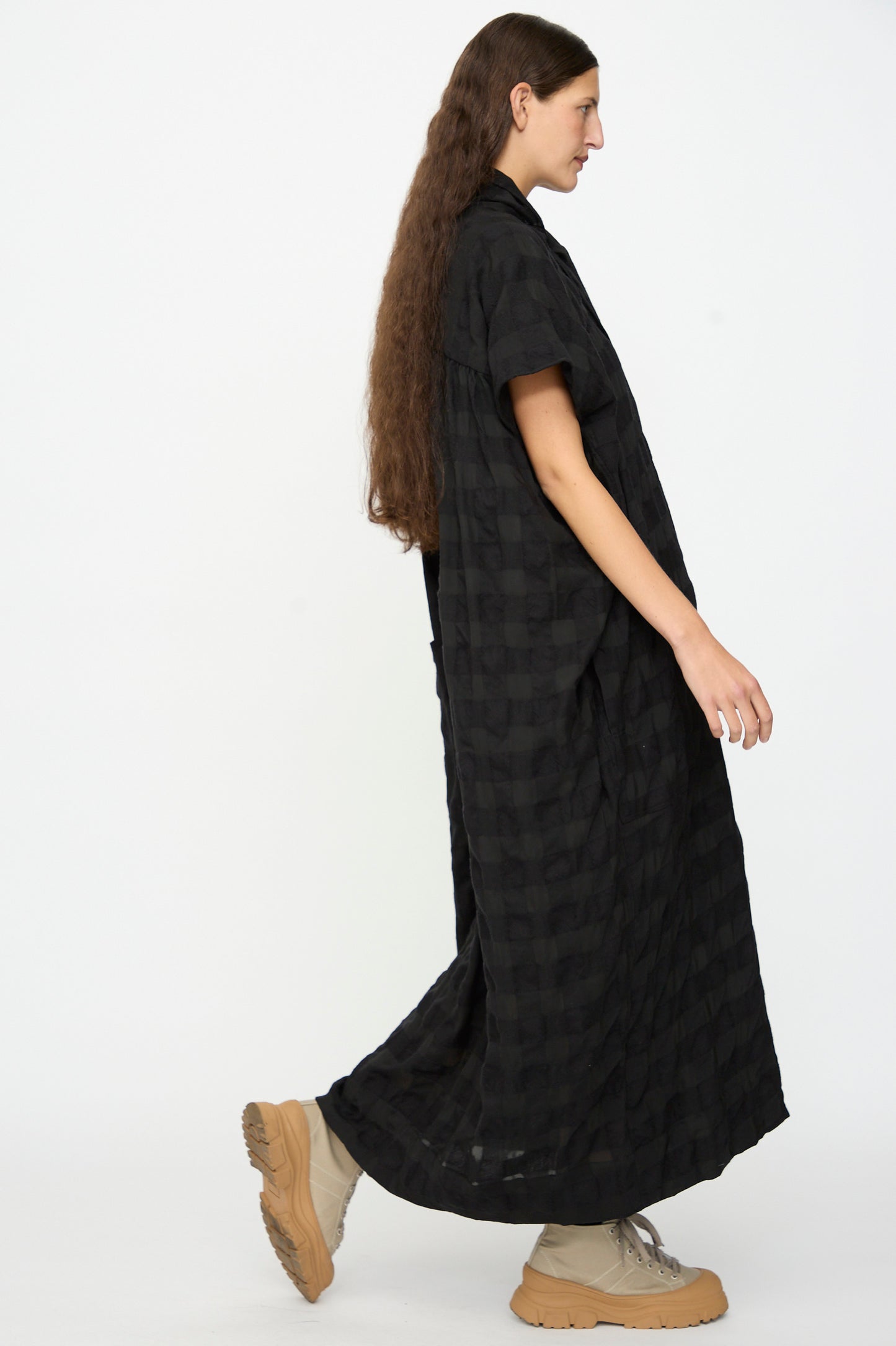 A person with long brown hair wears the SONO Dawn Dress in Black on Black—a loose, checkered wool-viscose blend maxi dress—paired with beige boots, walking in side profile against a plain white background.