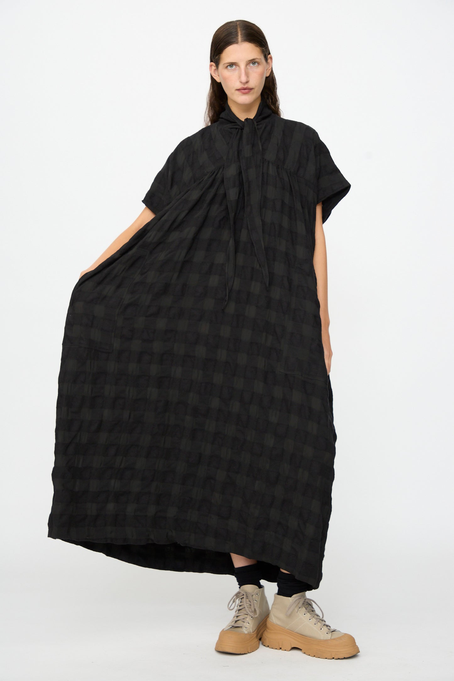 A person poses against a plain white background wearing the SONO Dawn Dress in Black on Black—a loose-fitting, short-sleeve maxi dress crafted from a wool-viscose blend with a black checkered pattern—paired with tan shoes.