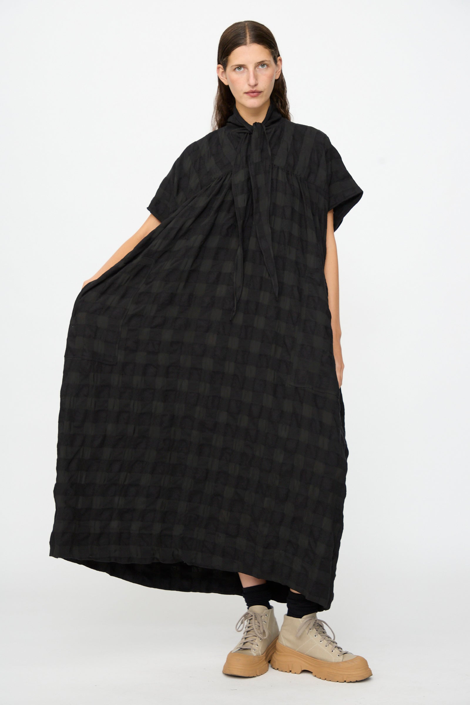 A person poses against a plain white background wearing the SONO Dawn Dress in Black on Black—a loose-fitting, short-sleeve maxi dress crafted from a wool-viscose blend with a black checkered pattern—paired with tan shoes.