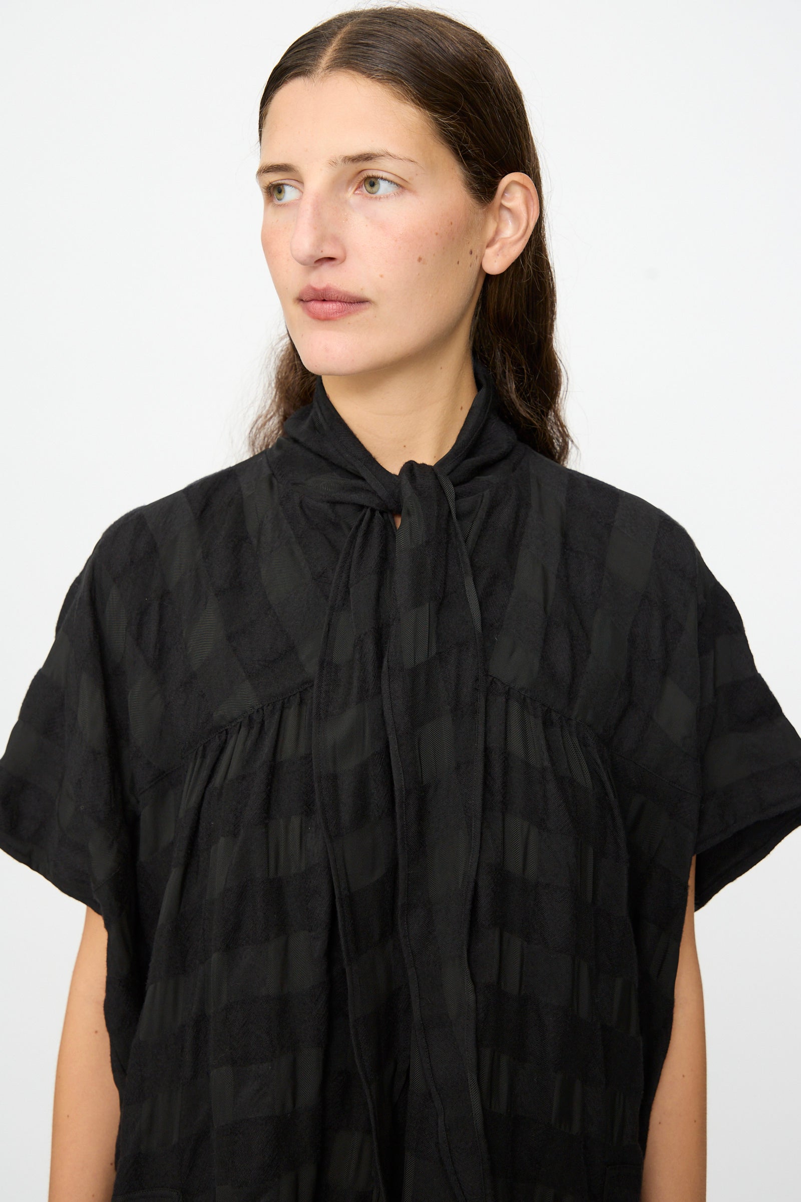 A person with long dark hair wears the SONO Dawn Dress in Black on Black, a short-sleeve, textured dress with a high necktie, crafted from a luxurious wool viscose blend, standing against a plain light background.