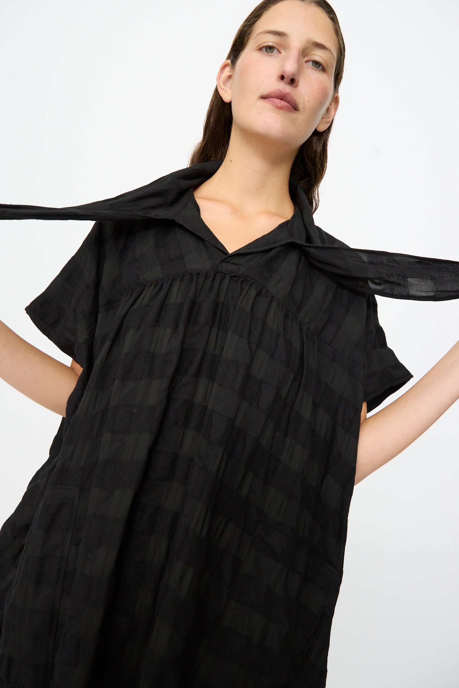 A person wearing the SONO Dawn Dress in Black on Black—a loose, short-sleeve maxi dress made from a luxurious wool viscose blend—with a matching scarf draped around their neck, standing against a plain white background. This elegant ensemble is made in France.