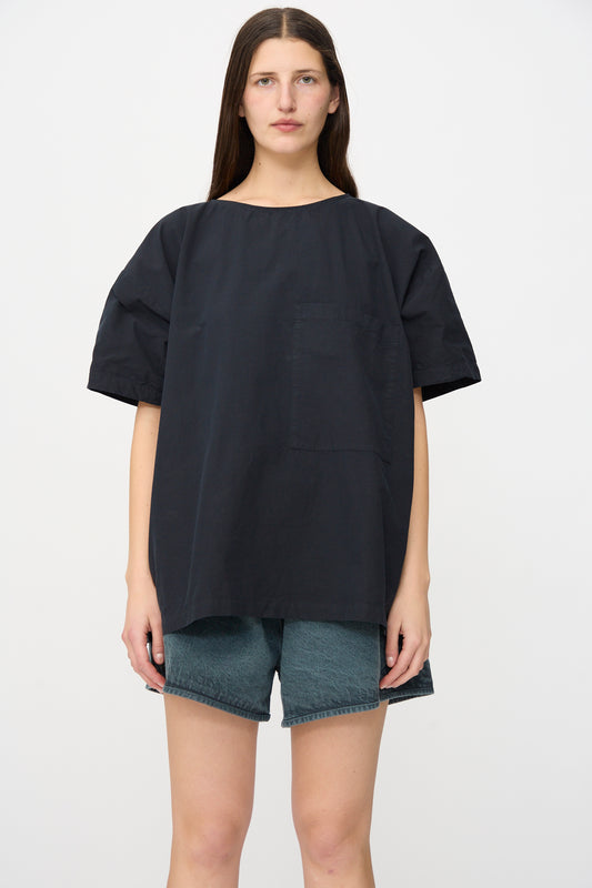 A person stands facing forward wearing the SONO Friedrich Tee in Off Black, an oversized fit boxy short sleeve shirt with a chest pocket, paired with greenish organic cotton denim shorts, set against a plain white background.