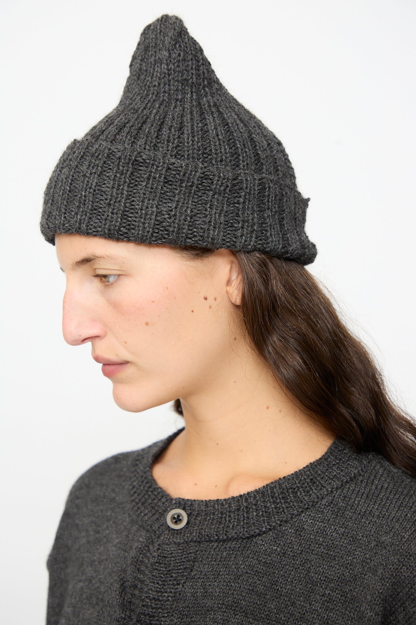 A person with long dark hair wears the SONO Gyu Beanie in Charcoal—a textured, pointed gray knit hat crafted in the UK from wool alpaca—along with a matching sweater, looking to the left against a plain background.