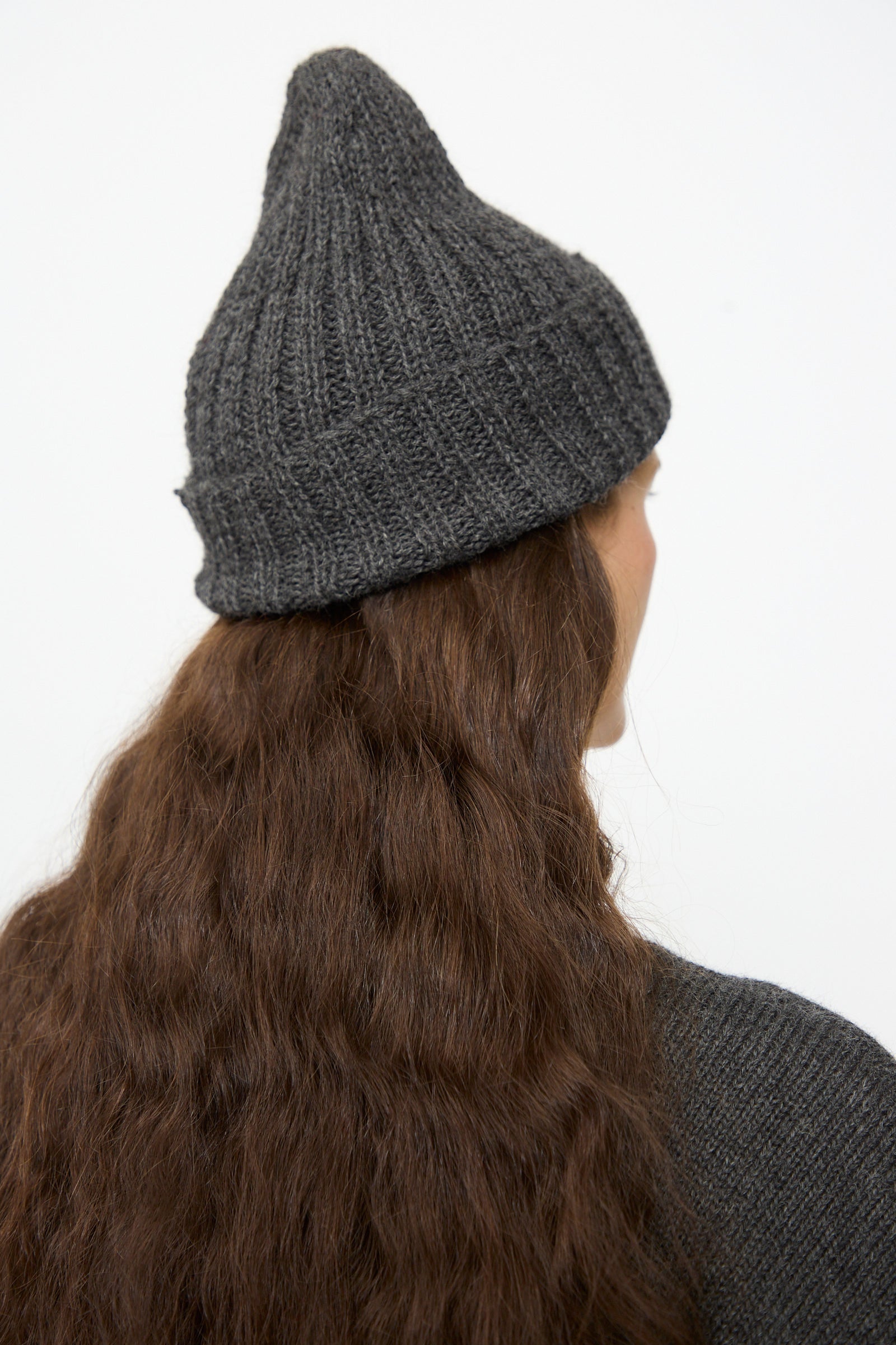 A person with long, wavy brown hair is shown from behind, wearing the SONO Gyu Beanie in Charcoal—a dark gray ribbed wool alpaca hat made in the UK—along with a matching sweater, set against a plain white background.