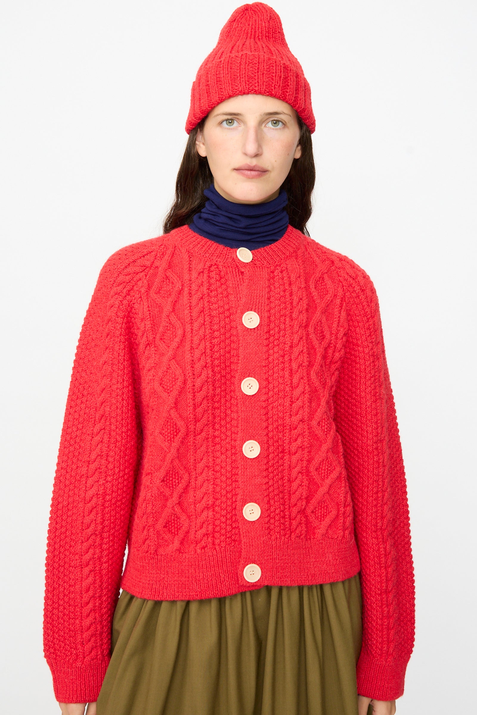 A person wearing the Gyu Beanie in Red by SONO—a bright red ribbed beanie made in the UK—paired with a matching red knit cardigan with buttons, a navy turtleneck, and an olive green skirt, stands against a plain white background.