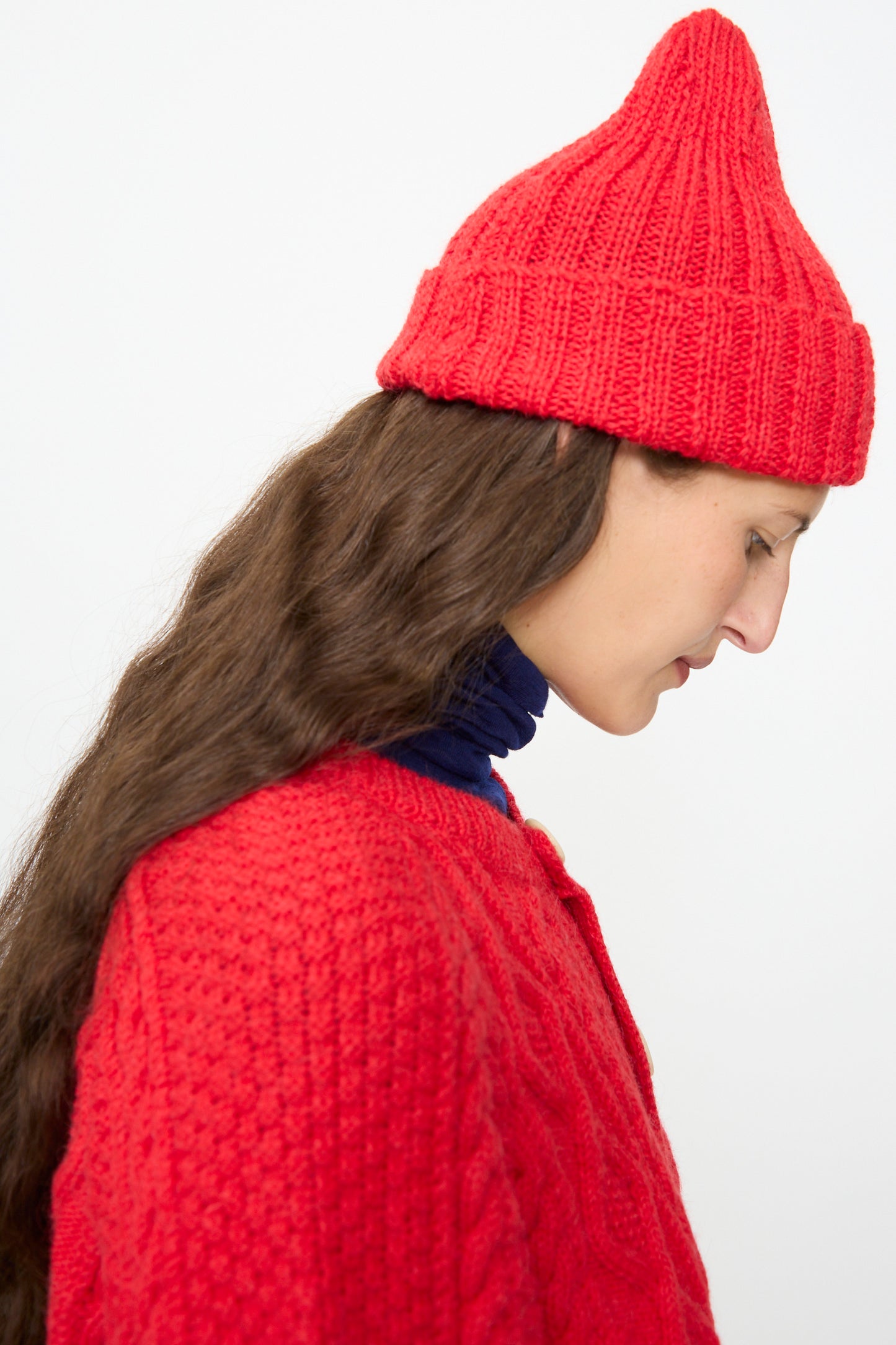 A woman in profile with long brown hair wears the SONO Gyu Beanie in Red and a red sweater layered over a blue turtleneck, set against a plain light background. The cozy wool alpaca beanie, crafted in the UK, by SONO, adds both warmth and style to her look.