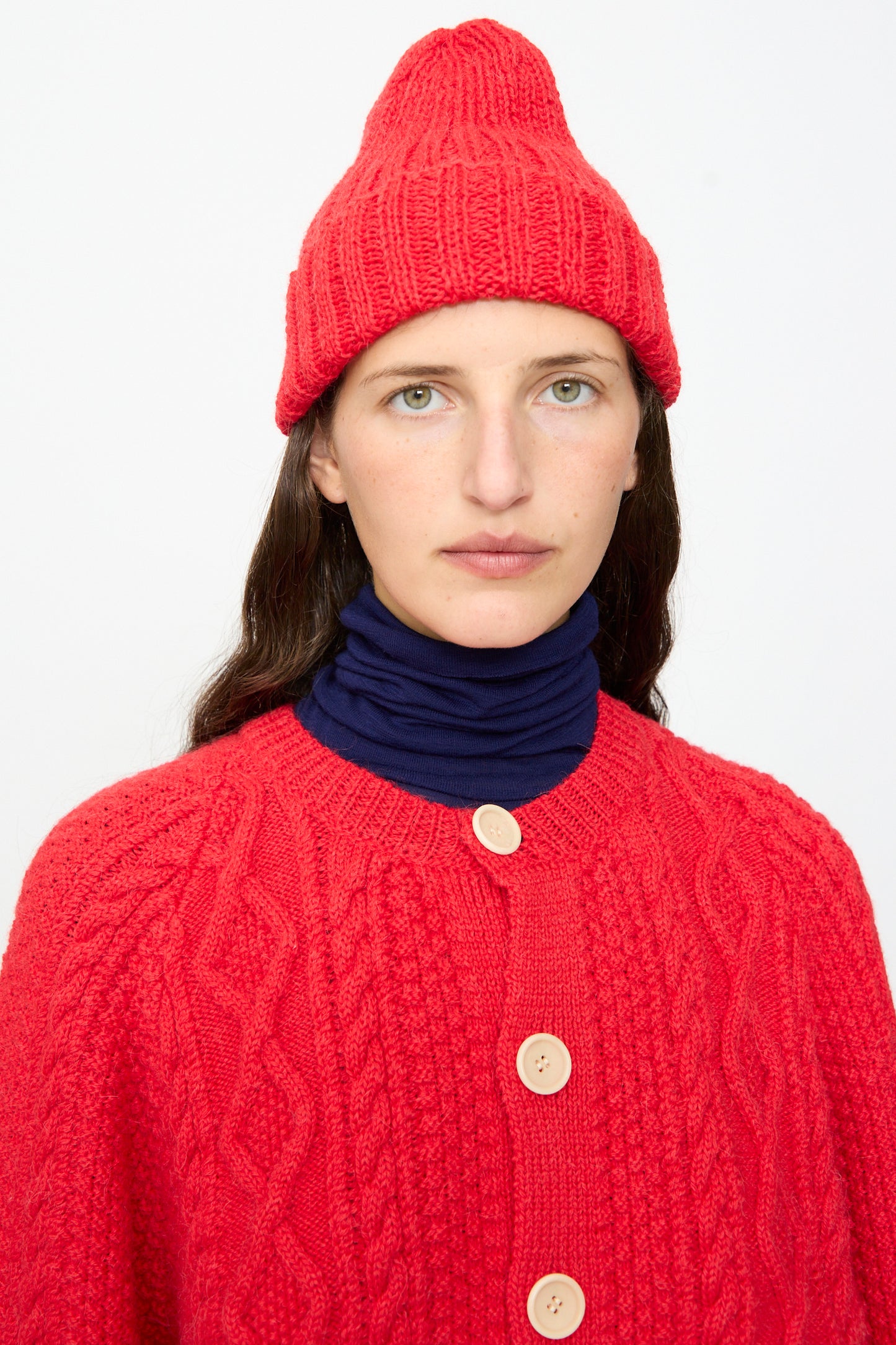A person wearing the SONO Gyu Beanie in Red and a red cardigan with large buttons over a navy turtleneck, facing the camera against a plain white background.