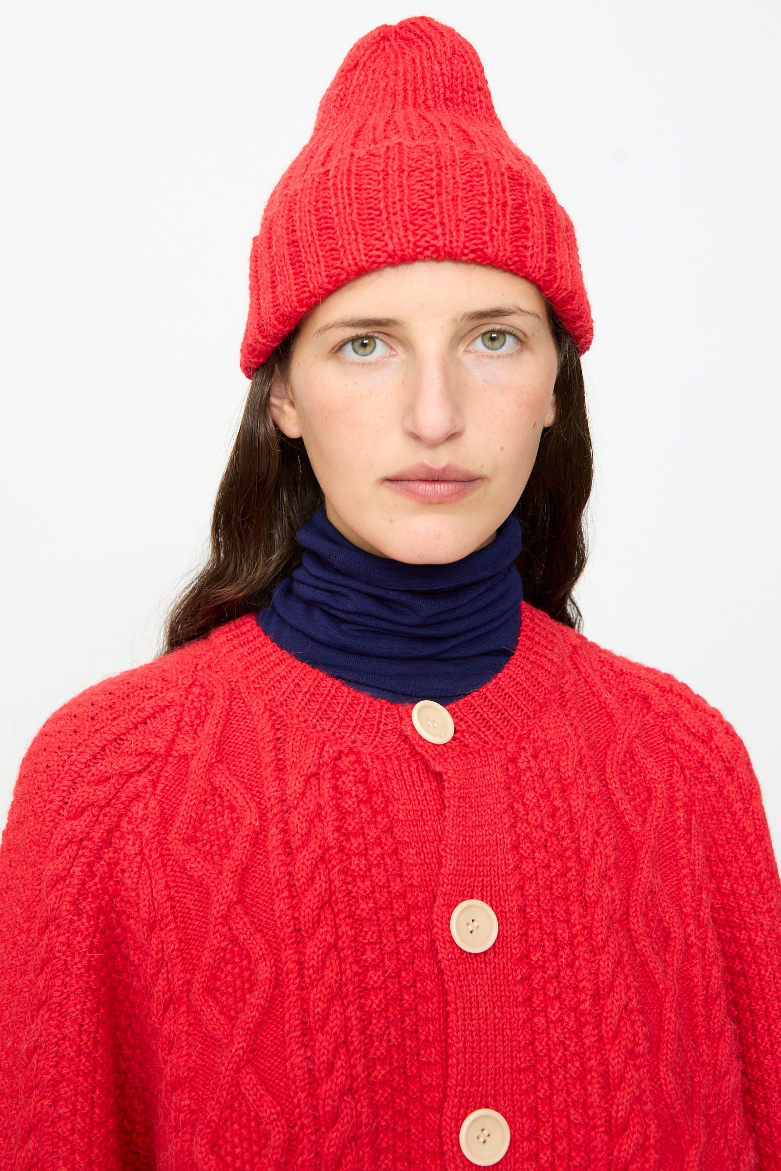 A person wearing the SONO Gyu Beanie in Red and a red cardigan with large buttons over a navy turtleneck, facing the camera against a plain white background.