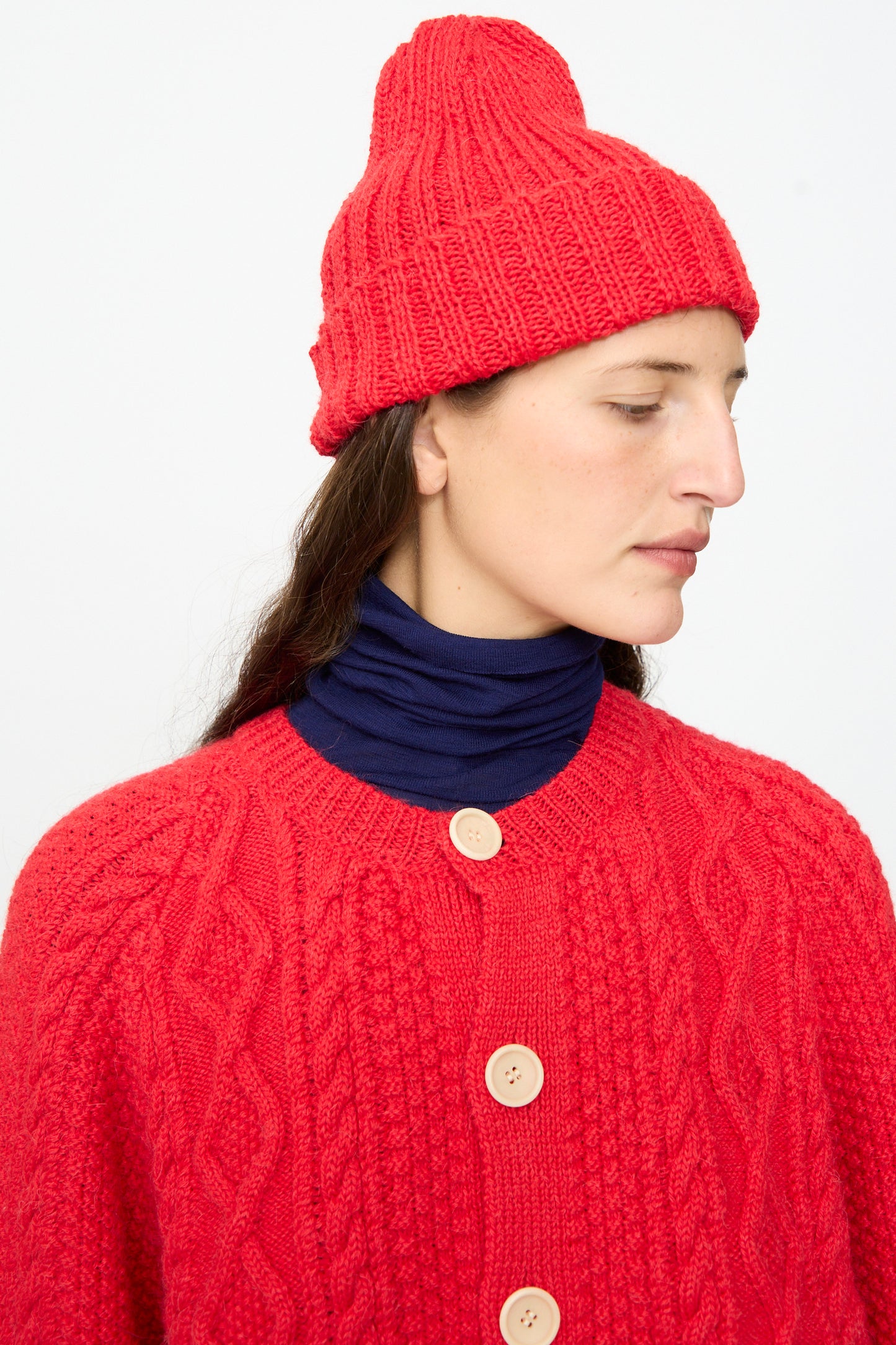 A person wearing the SONO Gyu Beanie in Red—crafted in the UK—paired with a matching red sweater featuring large buttons over a navy blue turtleneck, looks to the side against a plain white background.
