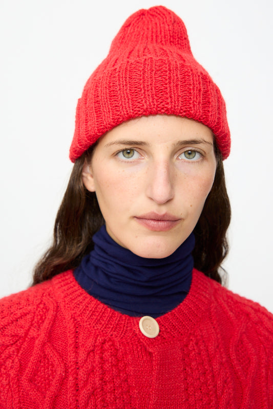 A person wearing the Gyu Beanie in Red by SONO and a sweater layered over a navy blue turtleneck looks directly at the camera against a plain background. Made in the UK from a wool-alpaca blend, this beanie adds cozy sophistication to their look.