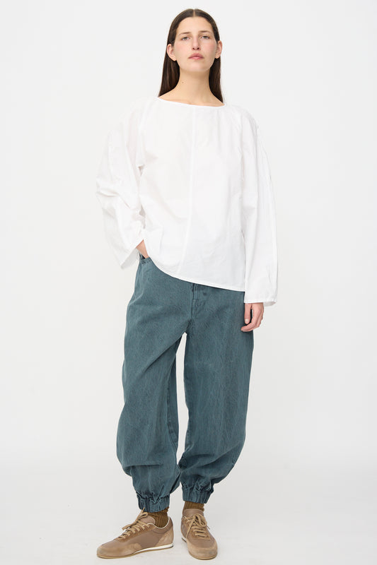 A person stands against a white background wearing a loose white long-sleeve shirt, SONO Jah Jeans in Bluestone with an elasticated waist and cinched ankles, and brown sneakers.