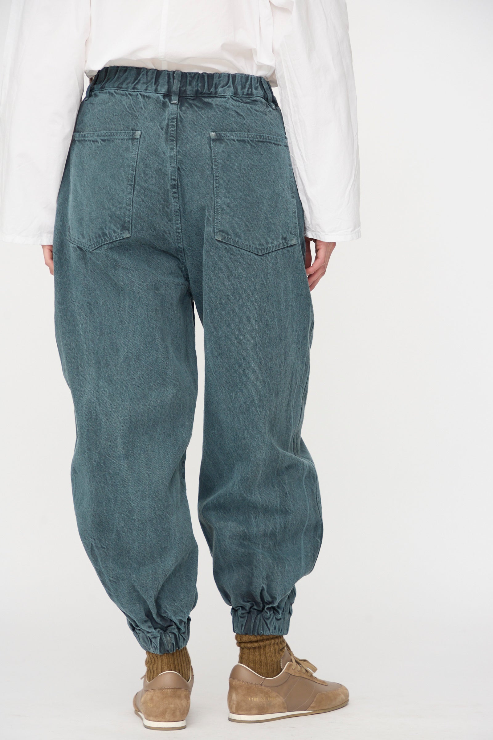 A person shown from the back wears SONO's Jah Jeans in Bluestone—relaxed, loose-fitting jeans with elasticated waist and cuffs—paired with a white long-sleeve shirt, tan shoes, and brown socks against a plain background.