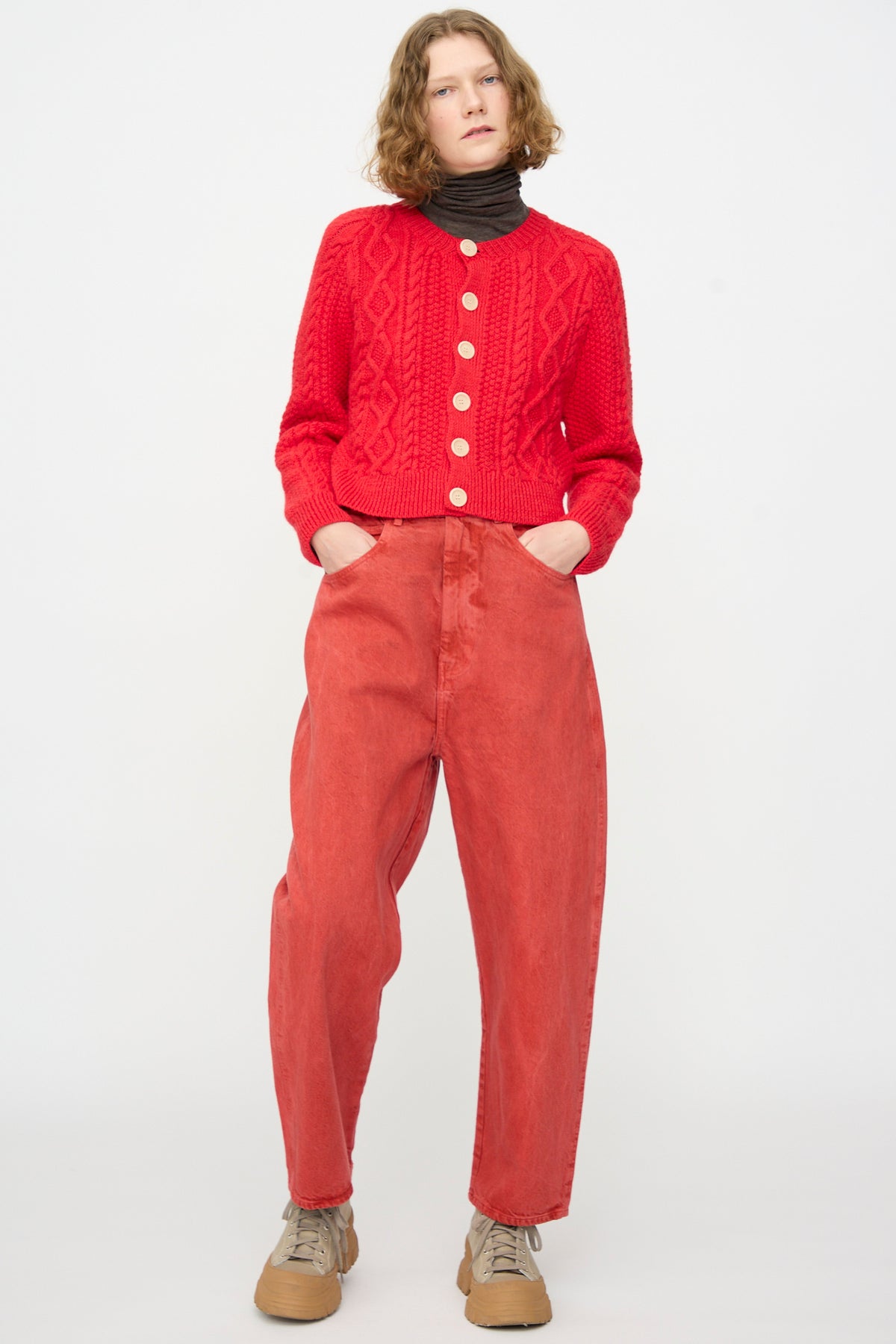 A person stands against a white background, wearing the SONO Jah Jeans in Red, a matching red knit cardigan, a black turtleneck, and tan sneakers with hands in their pockets.