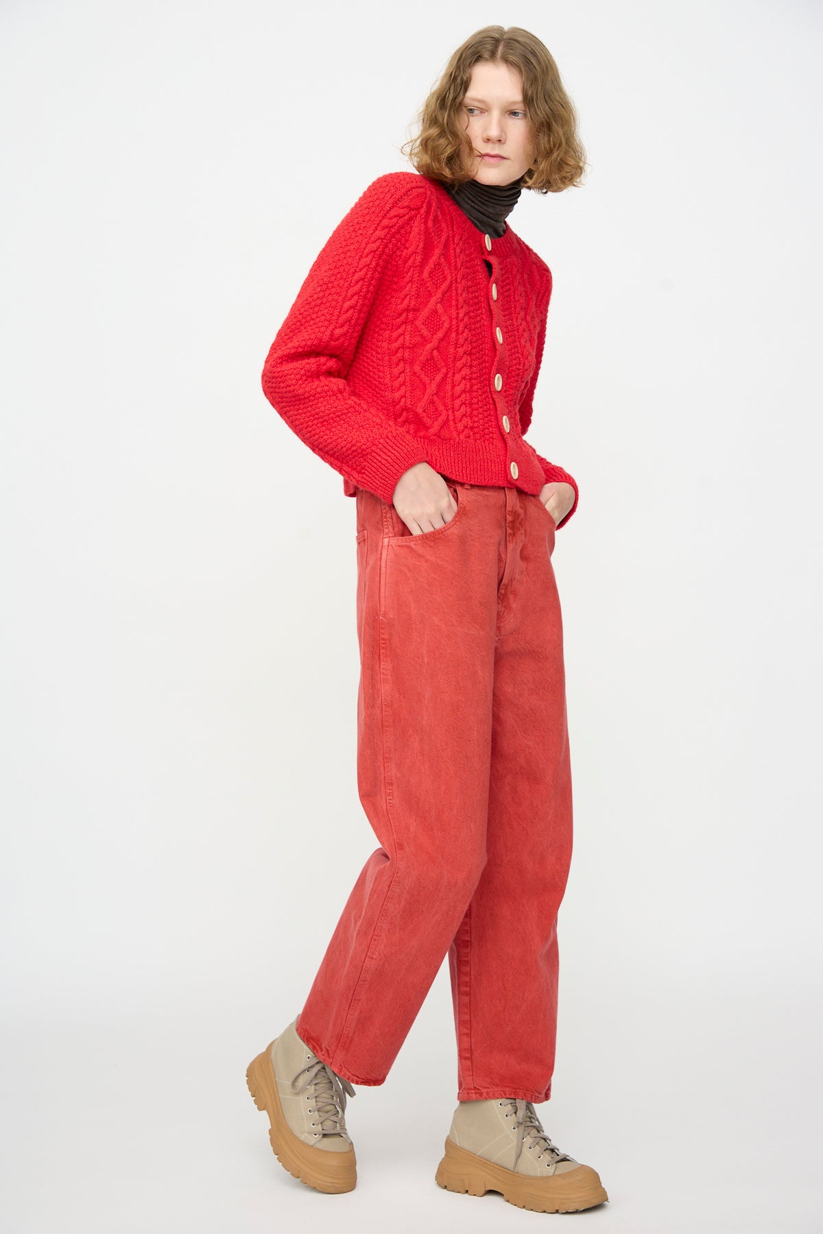 A person wearing SONO Jah Jeans in Red, a red cable-knit sweater, a black turtleneck, and beige boots stands against a plain white background.