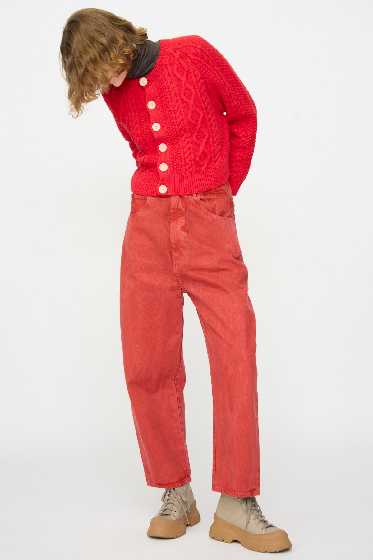 A person wearing a red knit sweater with large white buttons, SONO Jah Jeans in Red, beige boots, and a dark turtleneck stands against a plain white background while looking down.