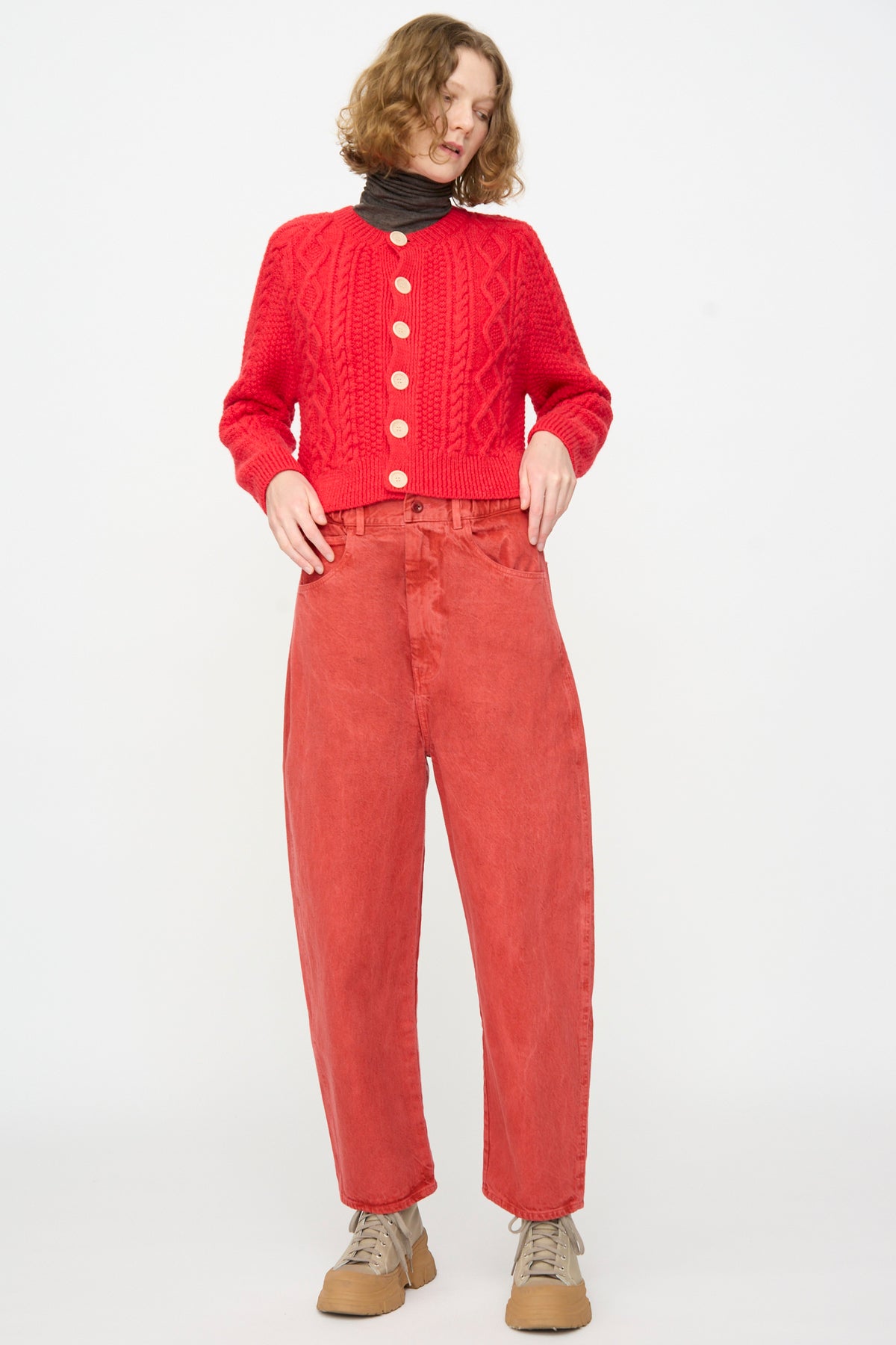 A person stands against a white background, wearing the SONO Jah Jeans in Red paired with a red cable knit cardigan, a dark turtleneck, and tan shoes.