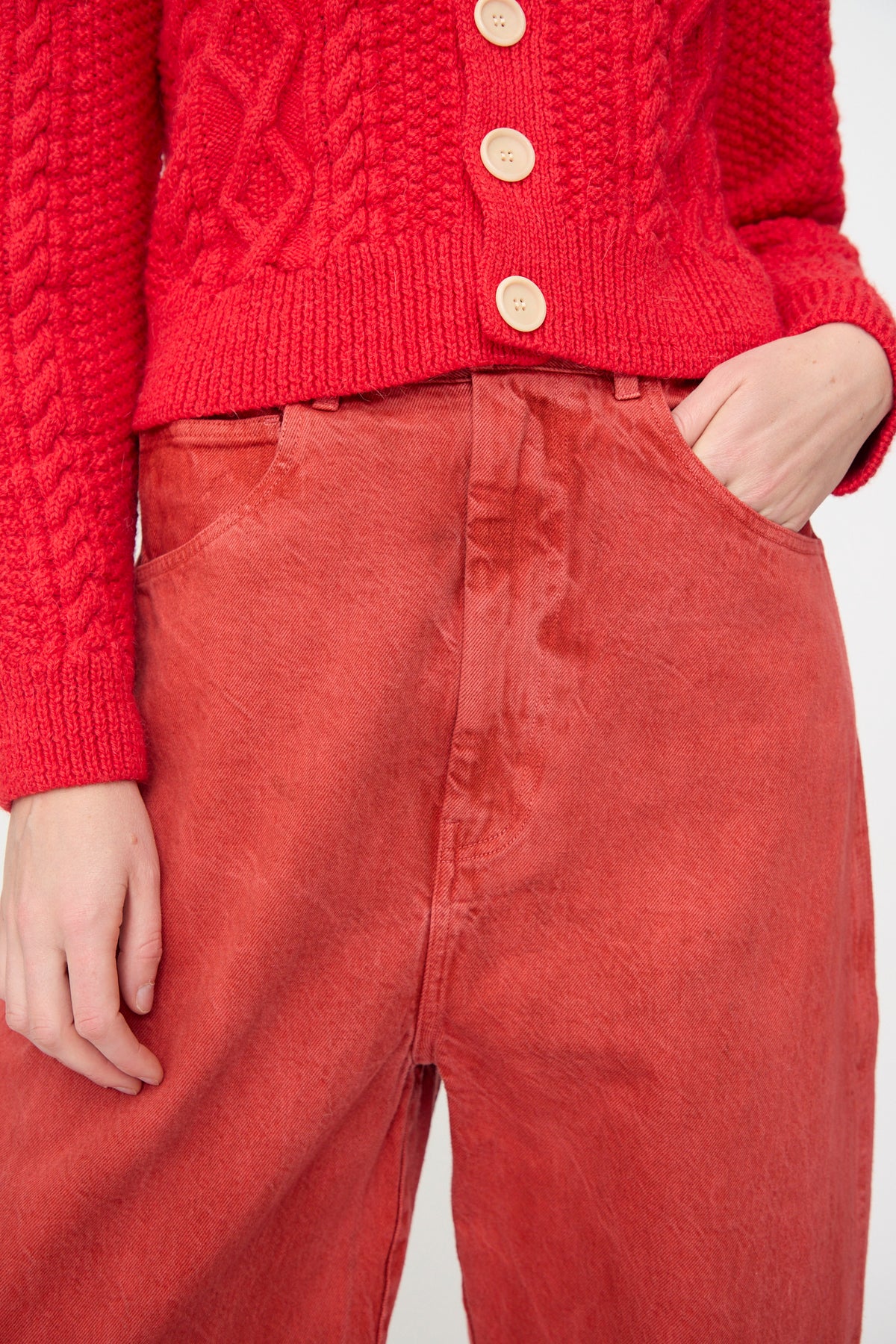 A person wearing SONO’s Jah Jeans in Red, styled with a textured red knit sweater with large buttons, stands with one hand in their pocket.