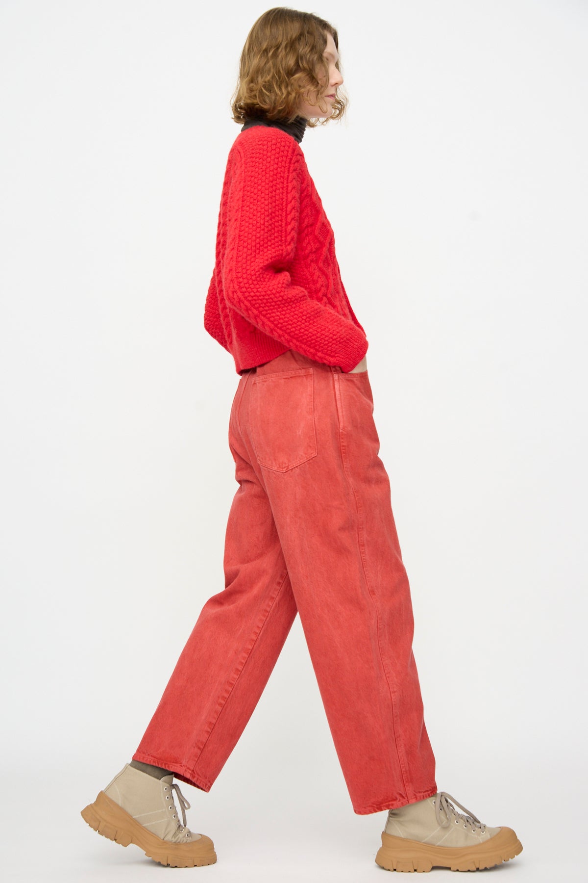 A person wearing SONO’s Jah Jeans in Red paired with a red sweater and beige boots walks to the left against a plain white background.