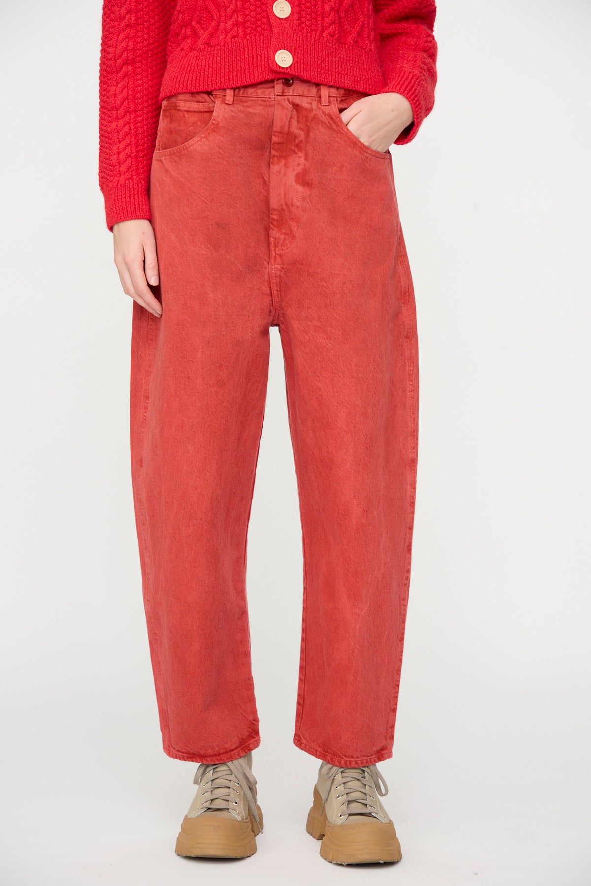 A person wearing SONO’s Jah Jeans in Red, styled with a red knit sweater and tan sneakers, stands against a plain white background. Only the lower torso and legs are visible.
