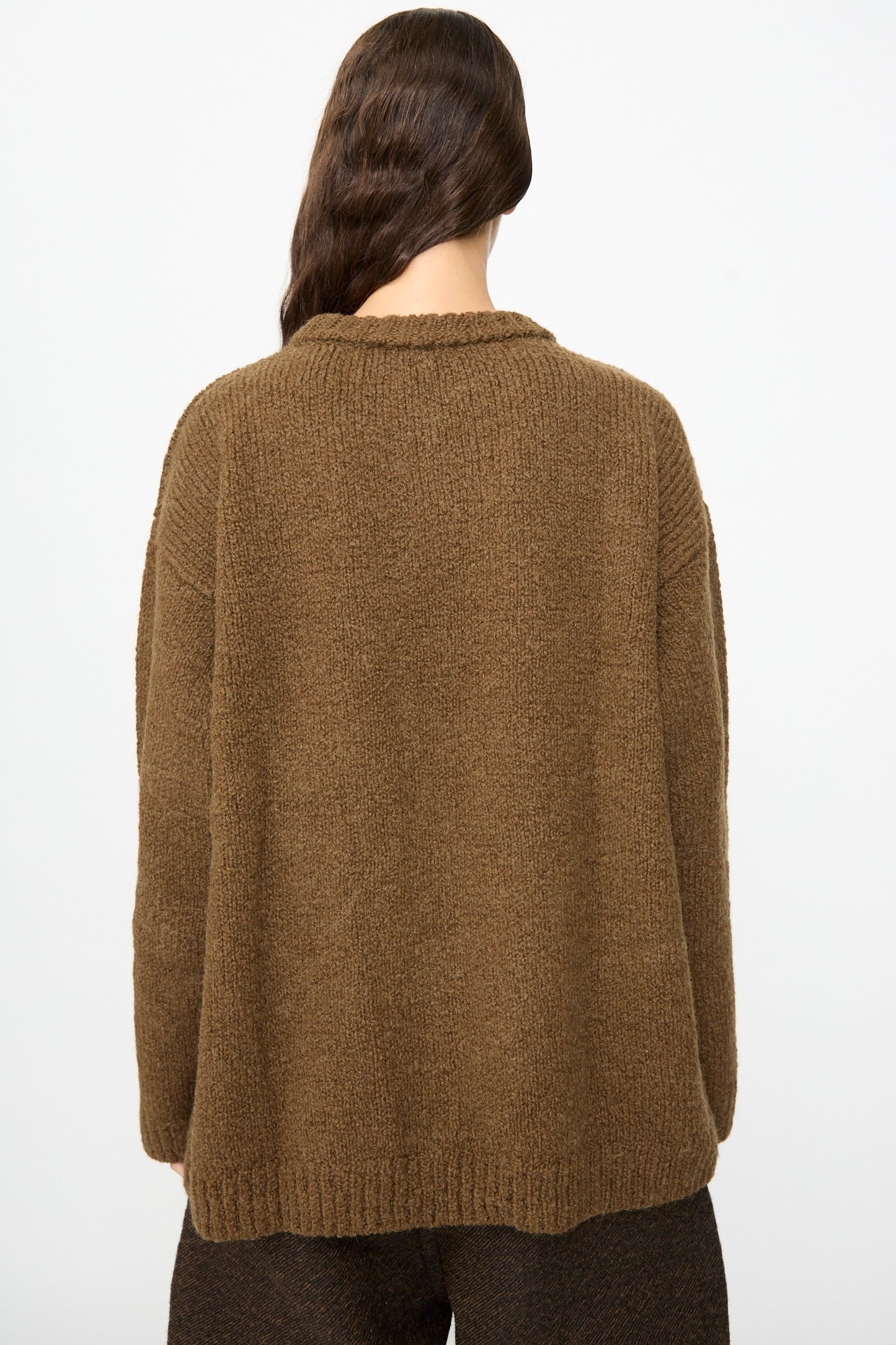 A person with long brown hair is shown from the back against a plain light background, wearing the SONO Jako Jumper in Kelp—an oversized pullover crafted from a soft wool-alpaca blend—paired with dark pants.