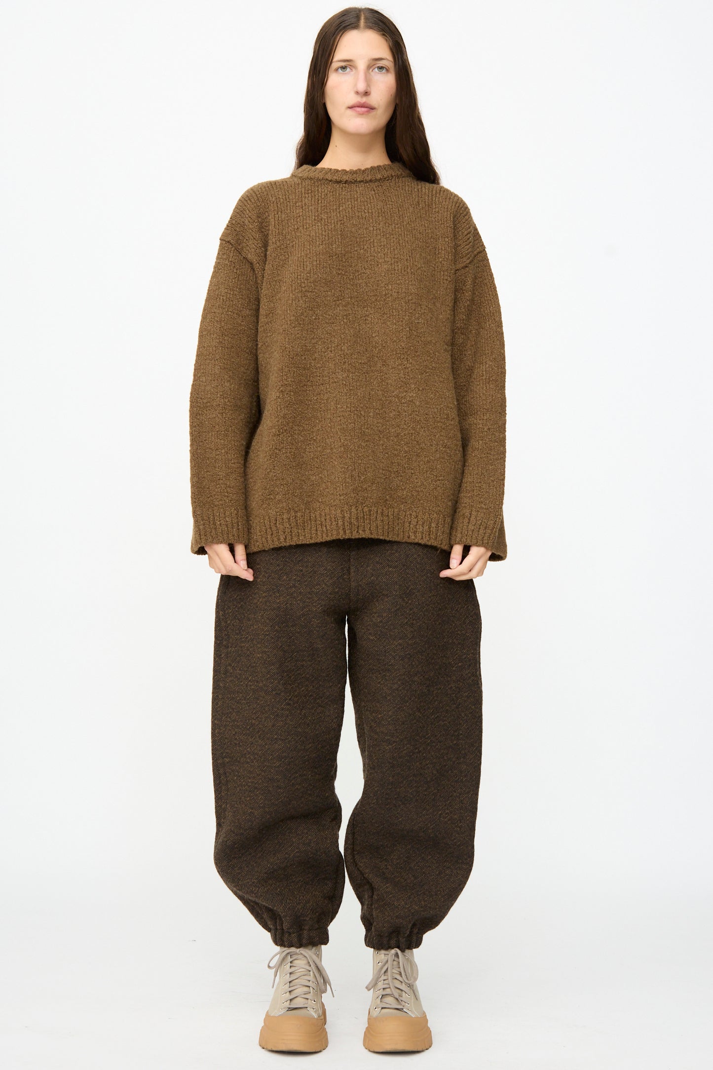 A person stands facing forward wearing the SONO Jako Jumper in Kelp, an oversized pullover crafted from a soft wool alpaca blend, paired with dark brown baggy pants with gathered cuffs and beige shoes, all set against a plain white background.