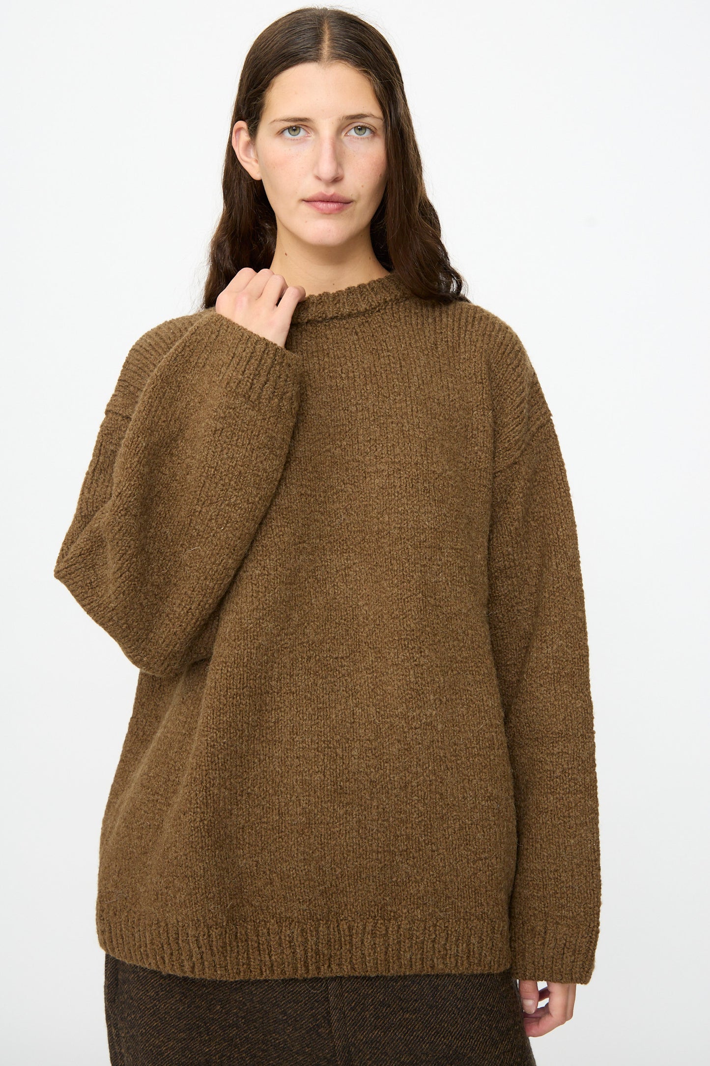 A person with long brown hair wears the SONO Jako Jumper in Kelp, an oversized pullover sweater crafted from a luxurious wool alpaca blend, paired with dark pants and standing against a plain white background.