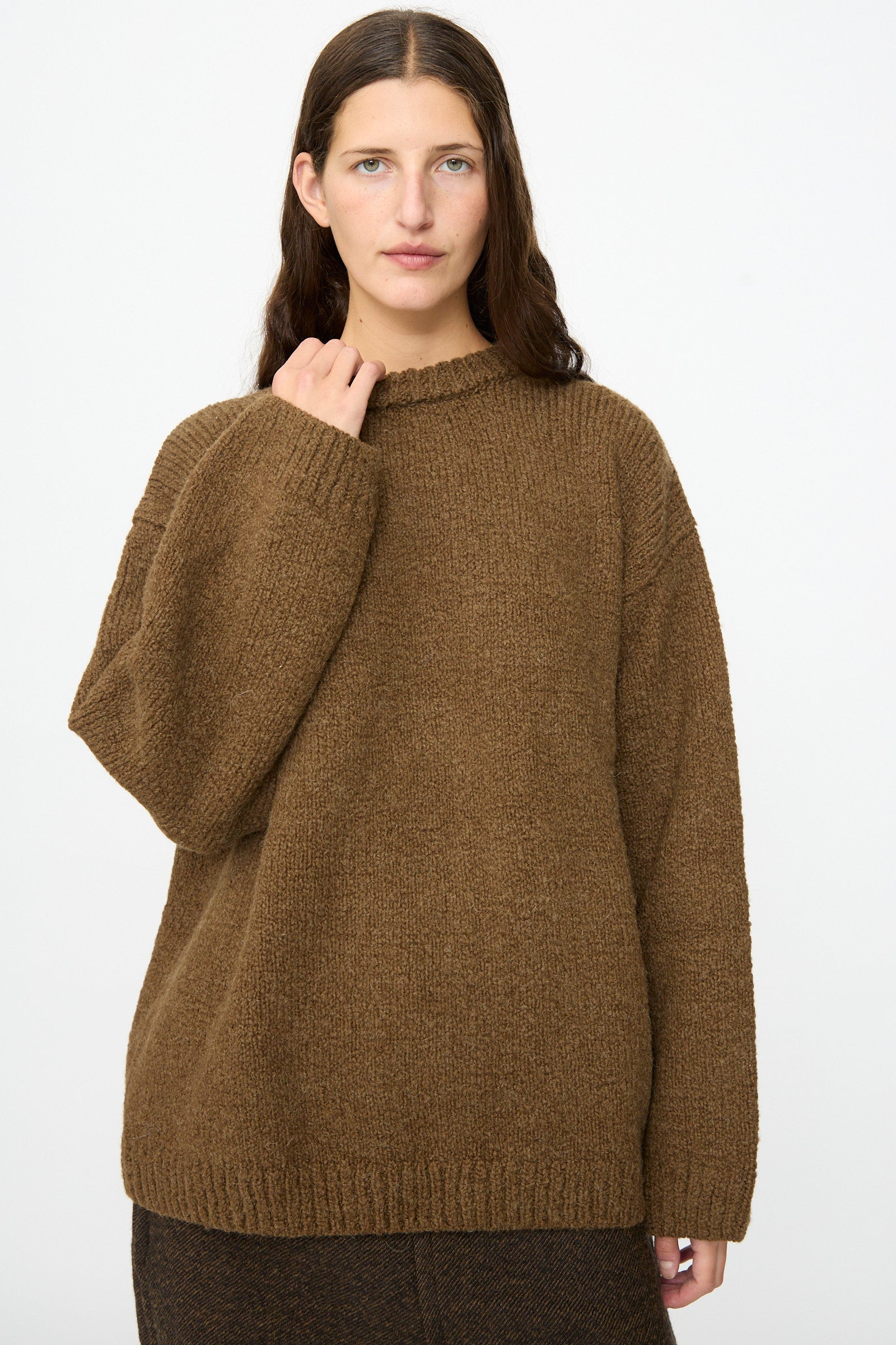 A person with long brown hair wears the SONO Jako Jumper in Kelp, an oversized pullover sweater crafted from a luxurious wool alpaca blend, paired with dark pants and standing against a plain white background.